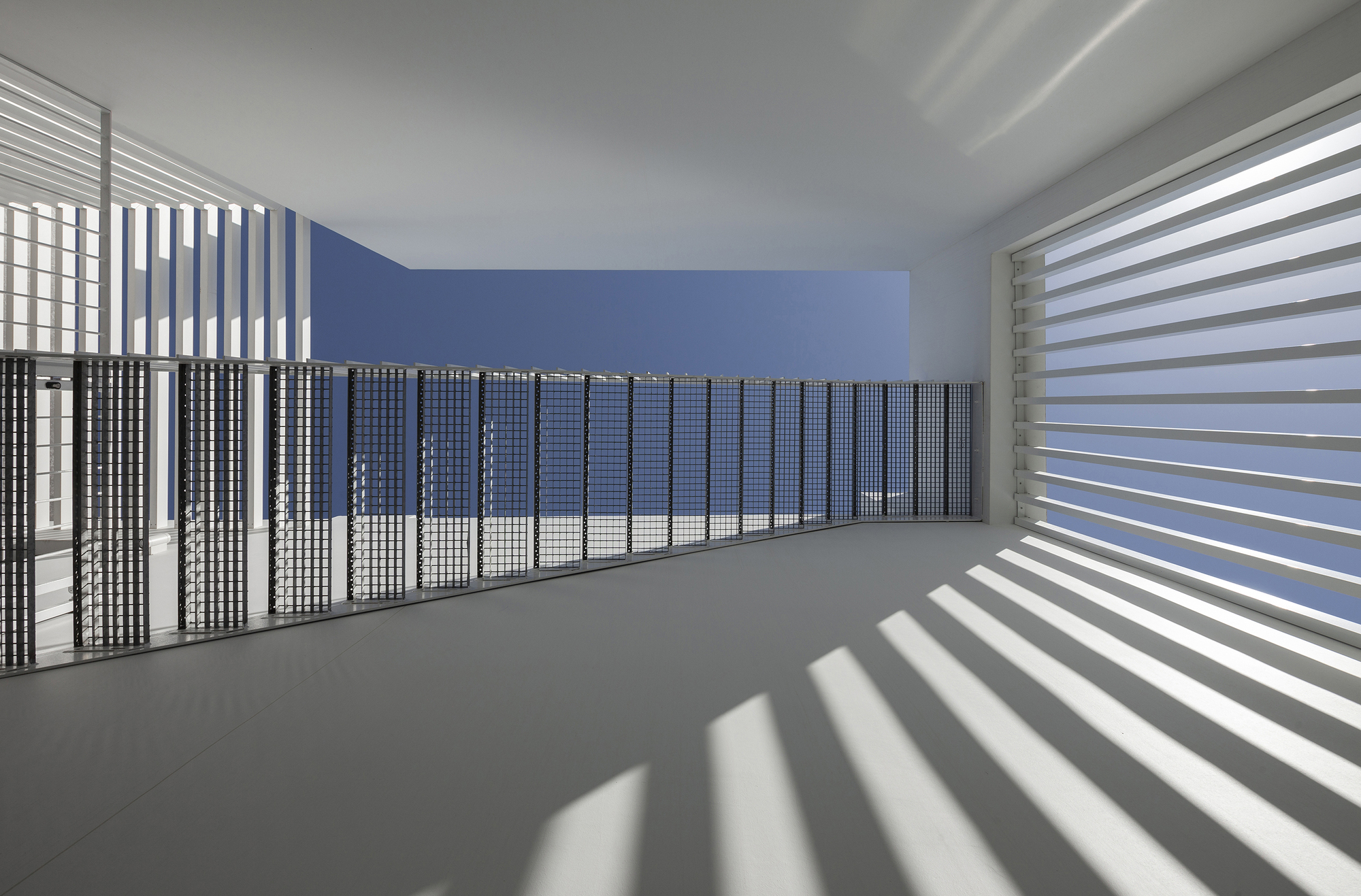 Upper level corridor with horizontal louvers casting striped shadows across white floors and blue wall