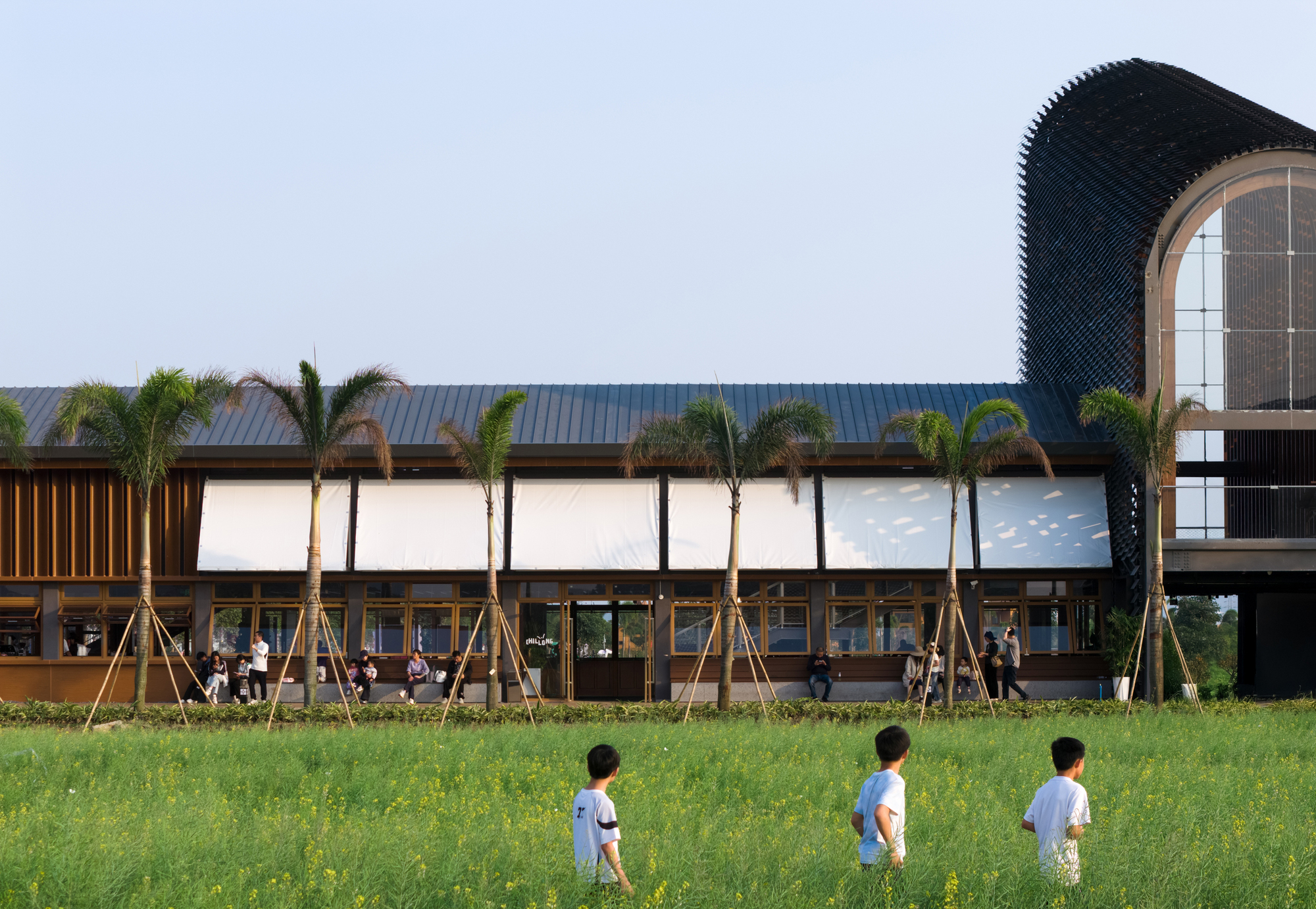 Facade with louvered panels and vertical timber screens beside a perforated brick tower and lawn