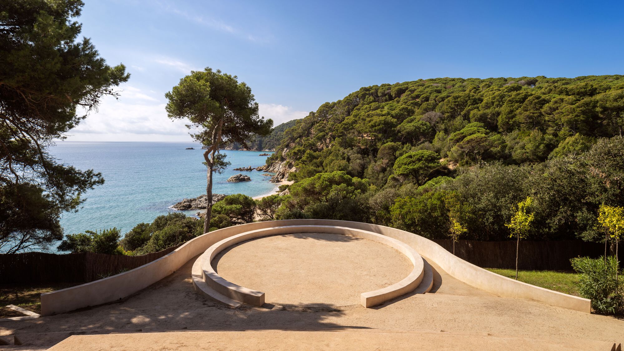 Curved amphitheatre with sand-colored terraces overlooking a forested coastal hillside and the sea