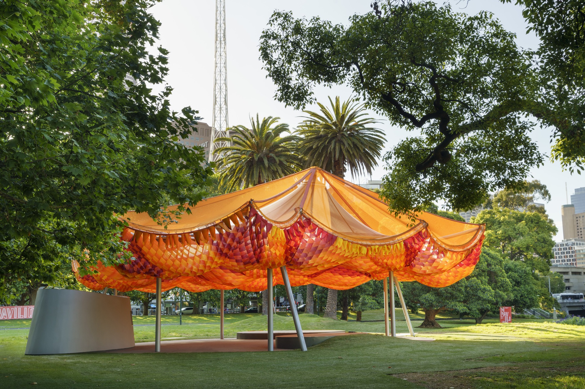 Tensile canopy with orange woven panels supported by angled columns in a park with palm trees