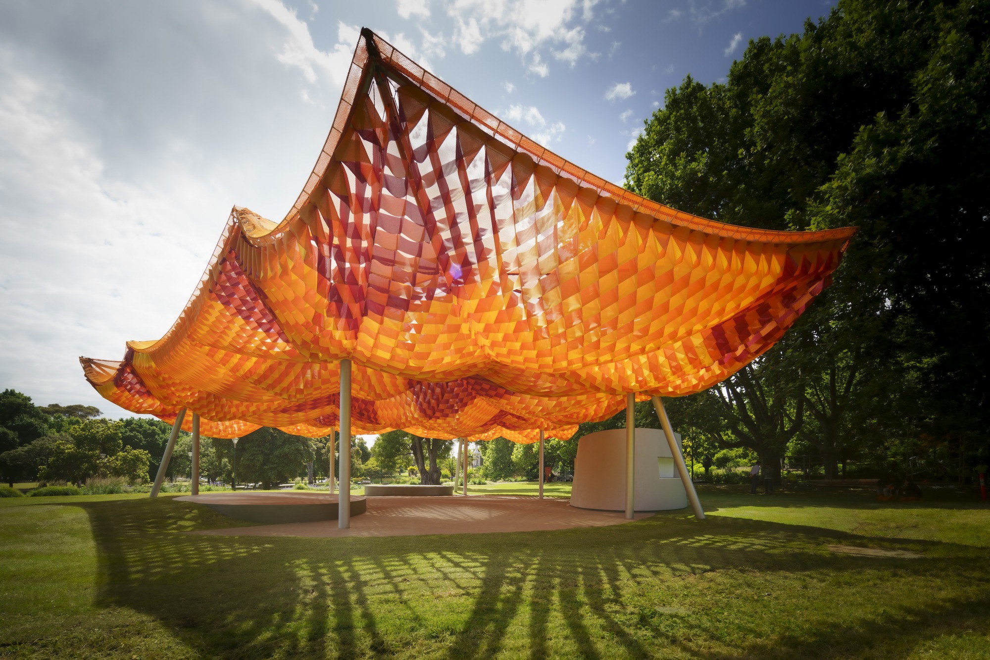 Side view of the scalloped canopy with woven panels creating dappled shadows on the paved ground