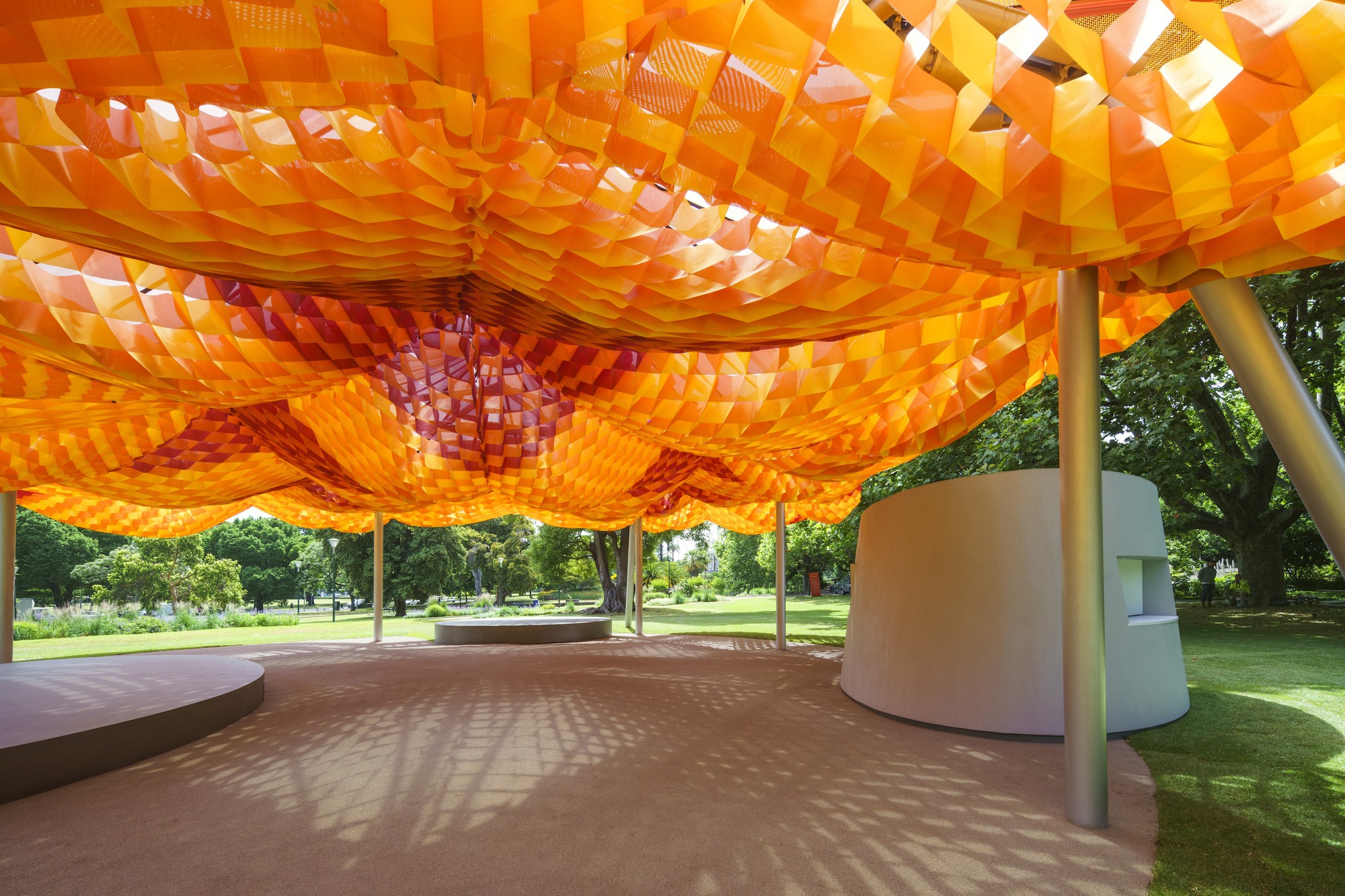 Interior view beneath the orange woven canopy showing light filtering through the geometric openings