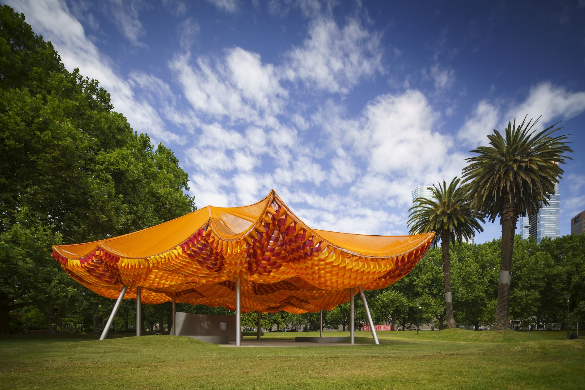 Long view of the tensile canopy structure set in a lawn with palm trees under blue sky