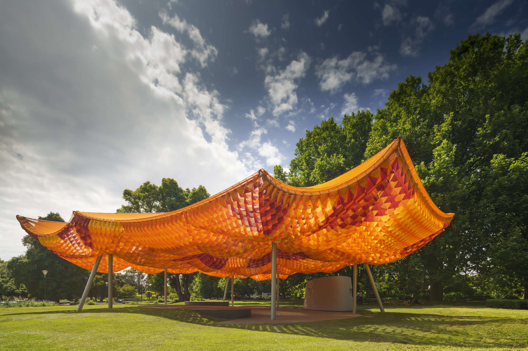 Woven orange canopy structure with upturned edges on white columns in a park under dramatic clouds