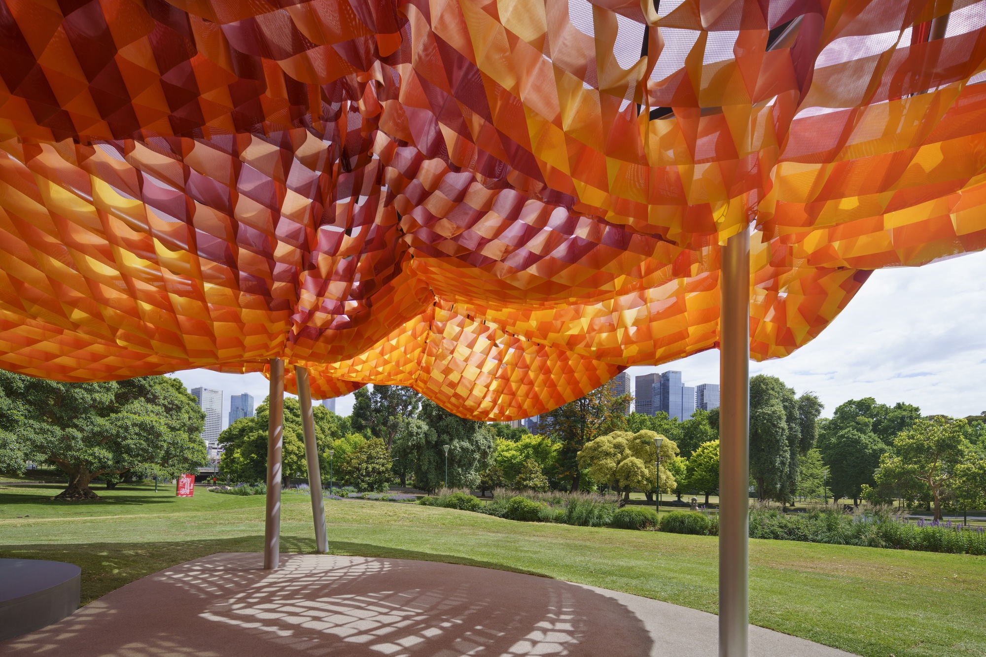 Underside view of the tessellated orange and red fabric canopy with park and skyline beyond