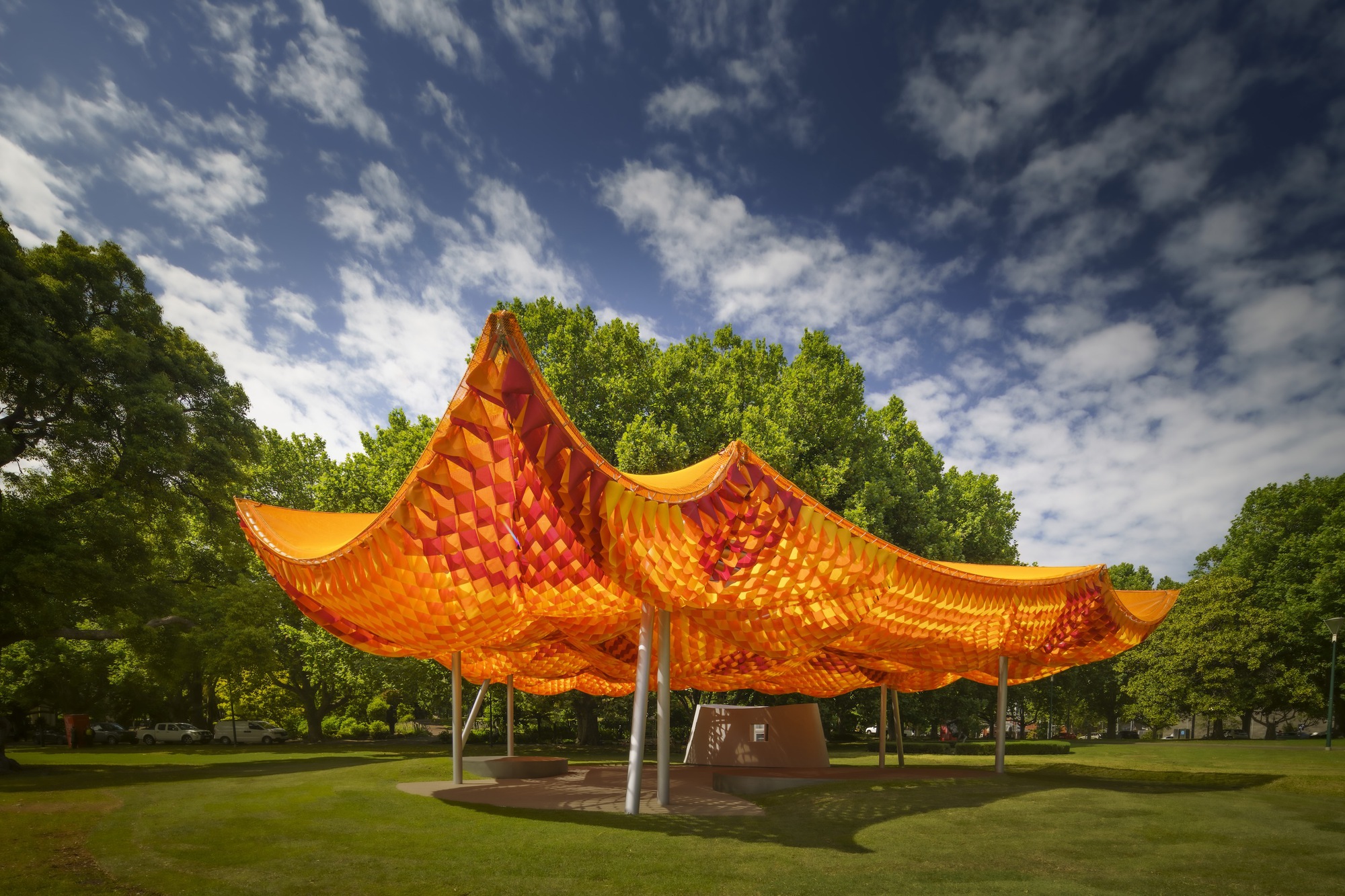Four-pointed woven canopy pavilion set in a lawn framed by mature trees under blue sky