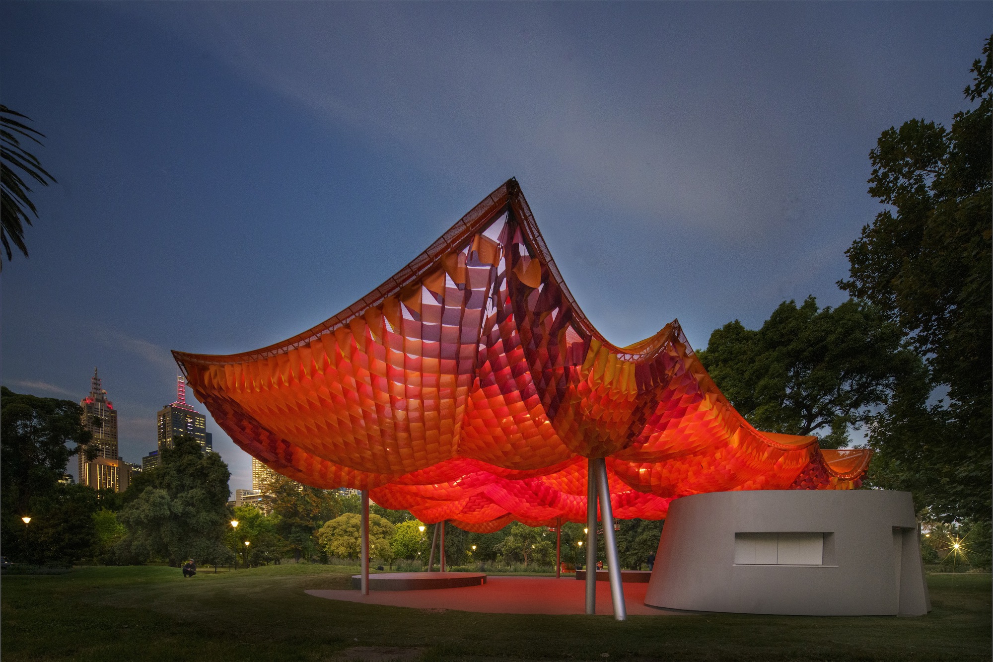 Dusk lighting transforms the canopy to glowing red above the cylindrical pavilion base in parkland