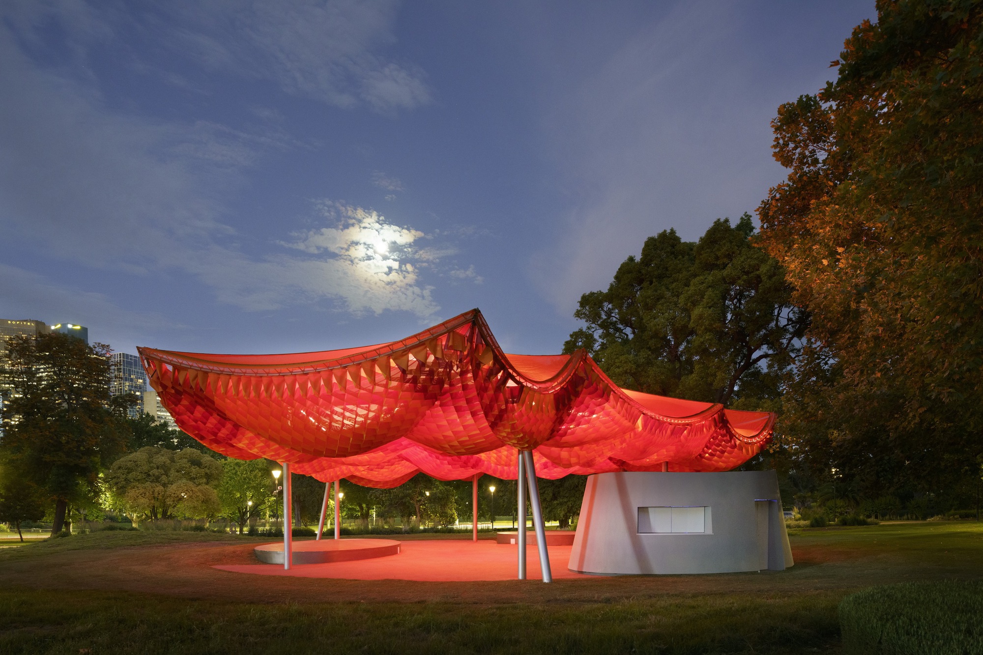 Pavilion with undulating red canopy on white columns in a park at twilight