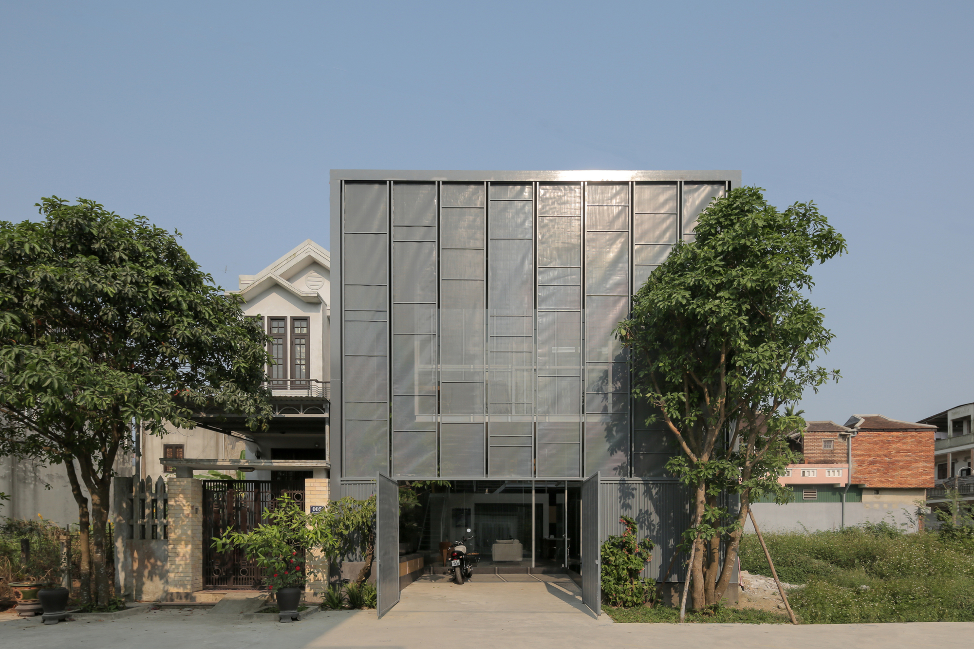 Street facade featuring a gridded metal screen wall flanked by mature trees and adjacent structures
