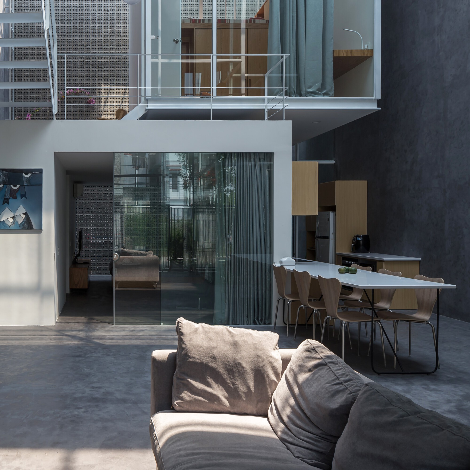 Double-height interior courtyard with glazed sliding doors, white steel balcony railing, and sunlight casting sharp shadows on polished concrete floor
