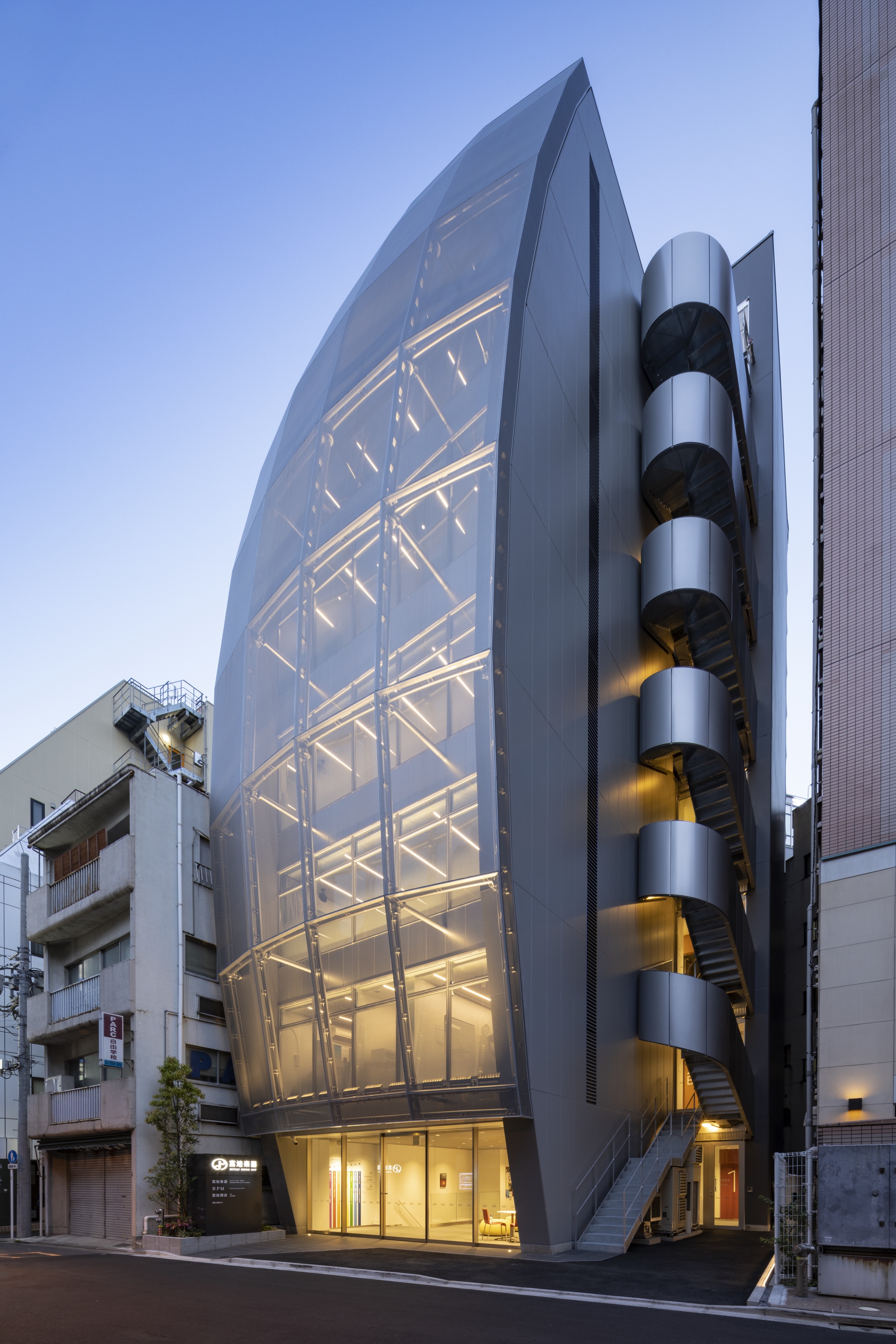 Street view of the faceted glass facade with illuminated structural frame and cylindrical stair tower at dusk