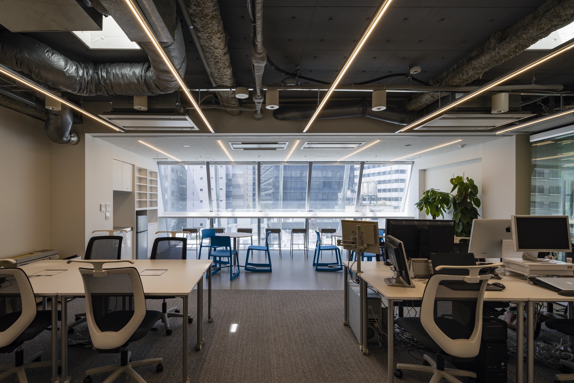 Open office floor with exposed ductwork, linear ceiling lights, and floor-to-ceiling windows overlooking neighboring buildings