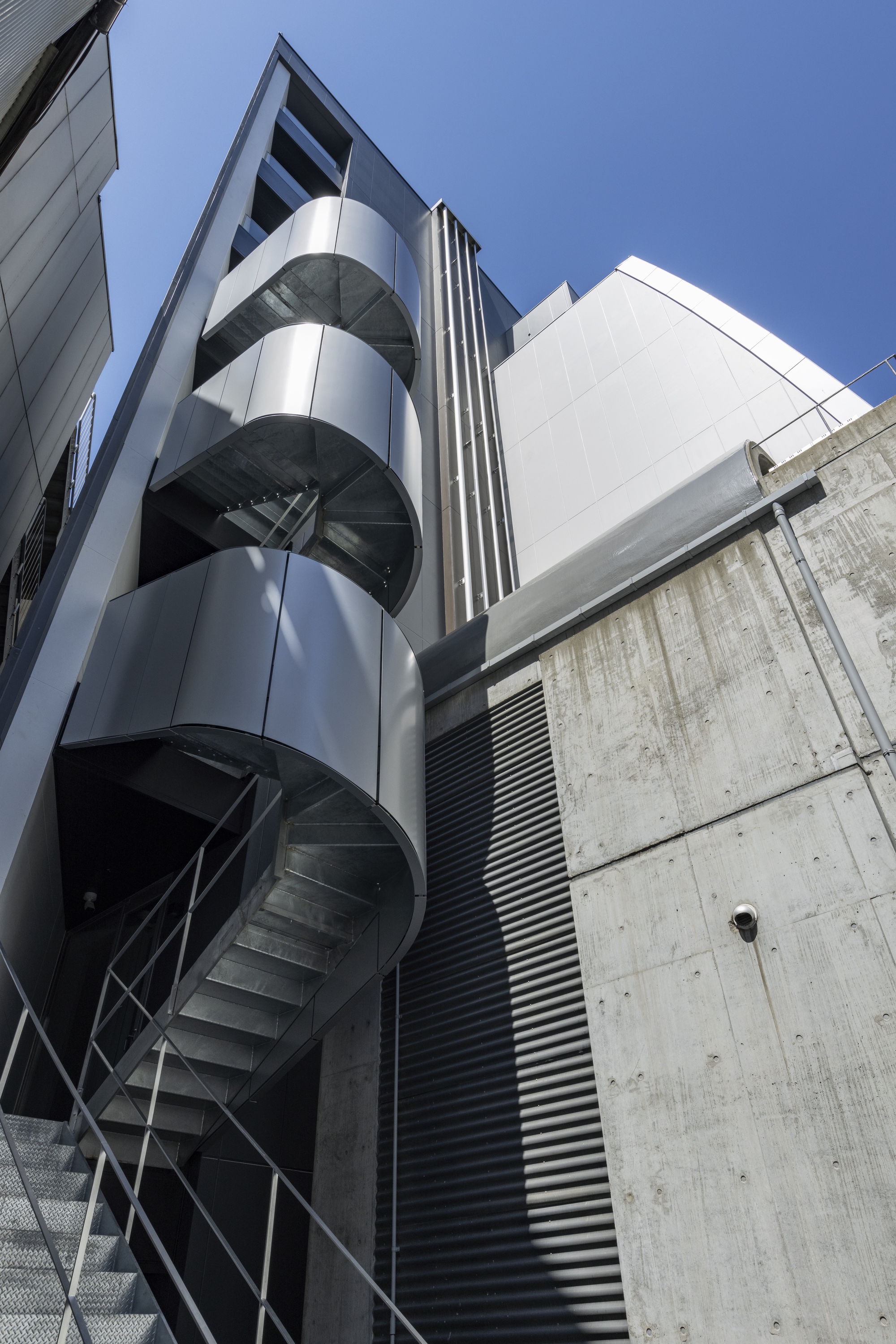 Exterior spiral staircase wrapping around a cylindrical glass tower beside a concrete wall