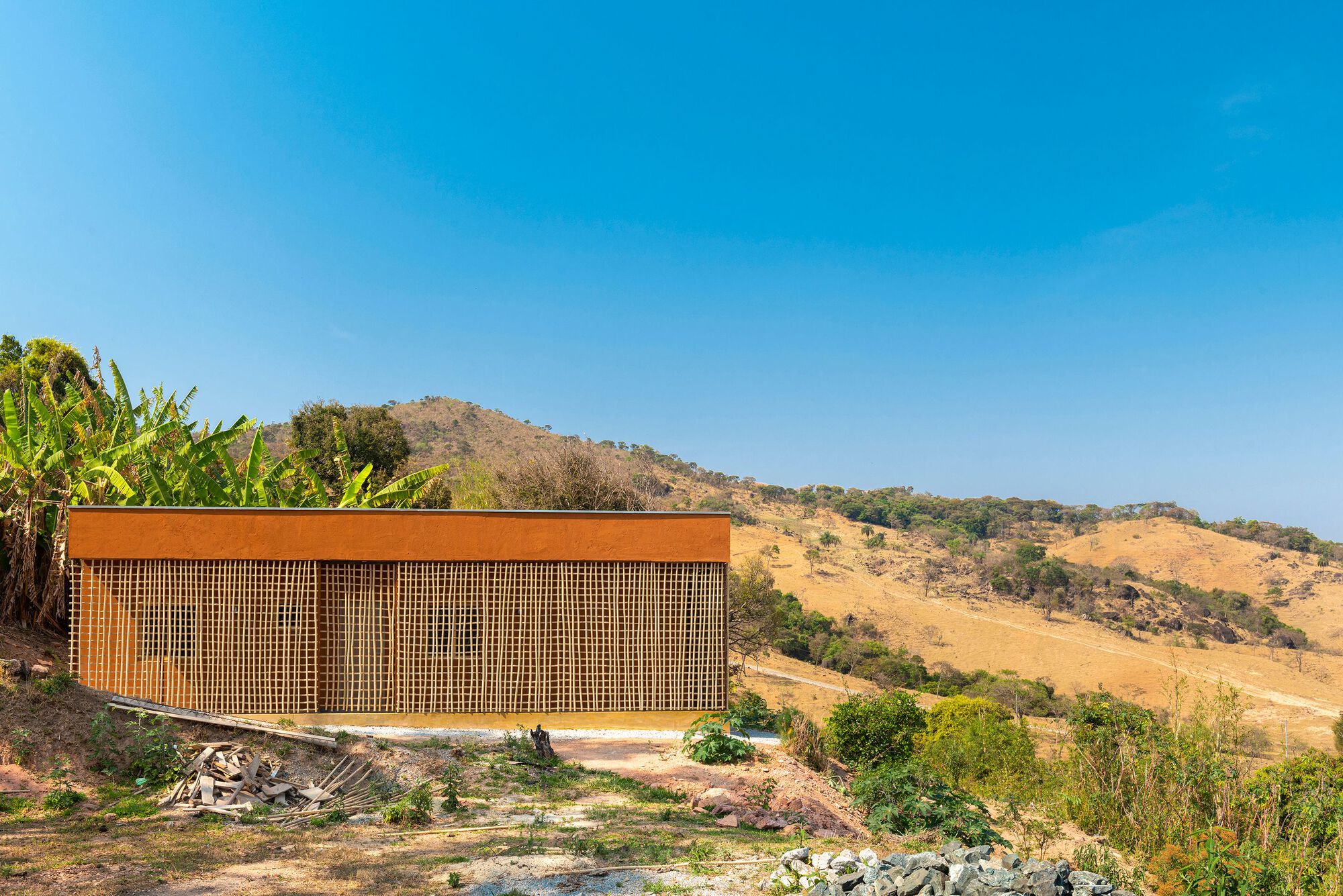 Elevated rectangular volume with timber lattice screen facade set against a dry hillside with banana plants