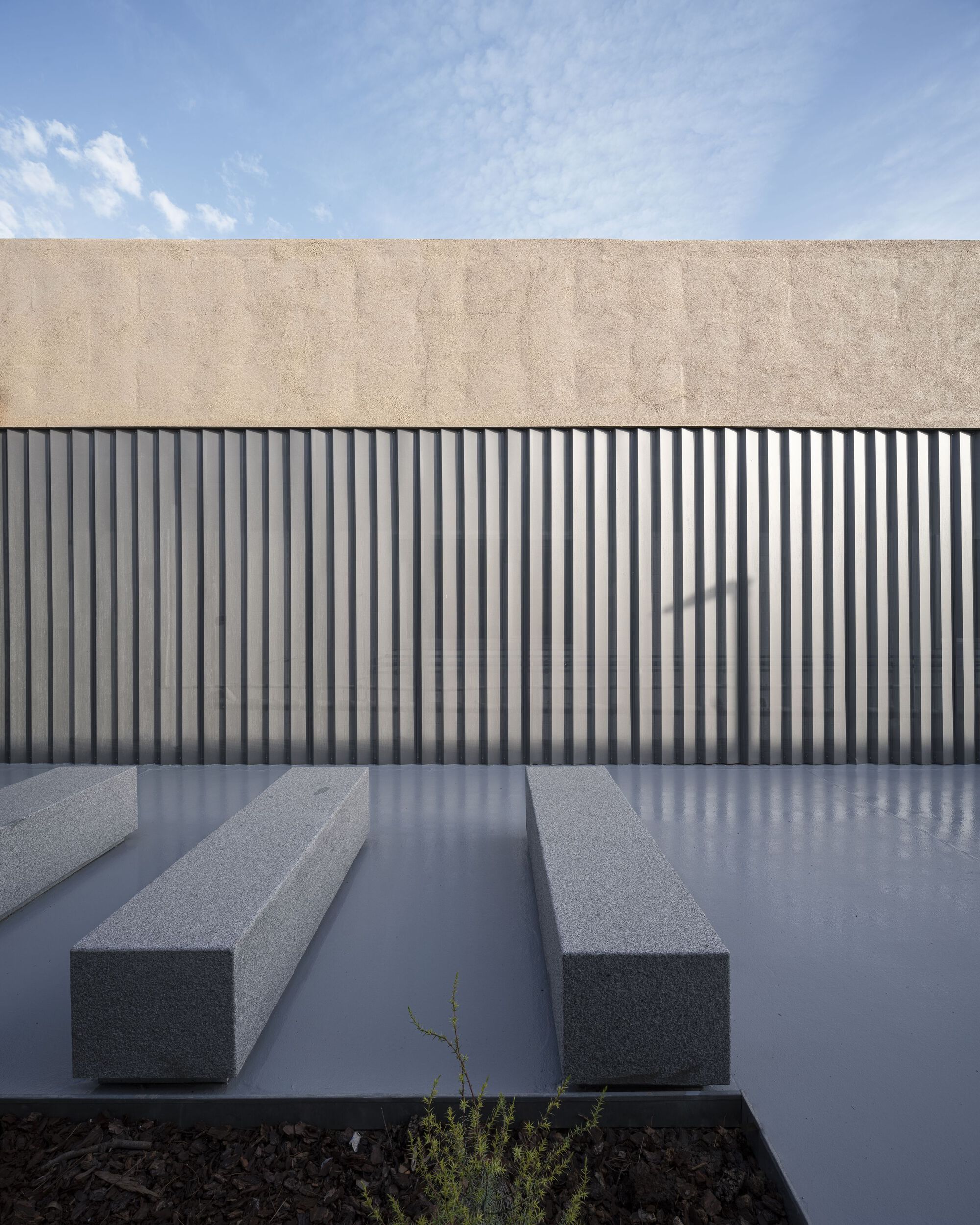 Courtyard with grey concrete benches and vertical metal siding below a beige plaster parapet under blue sky