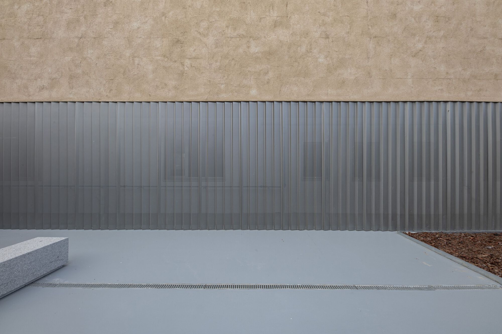 Close-up of vertical corrugated metal facade meeting concrete paving with linear drainage channel and planted bed
