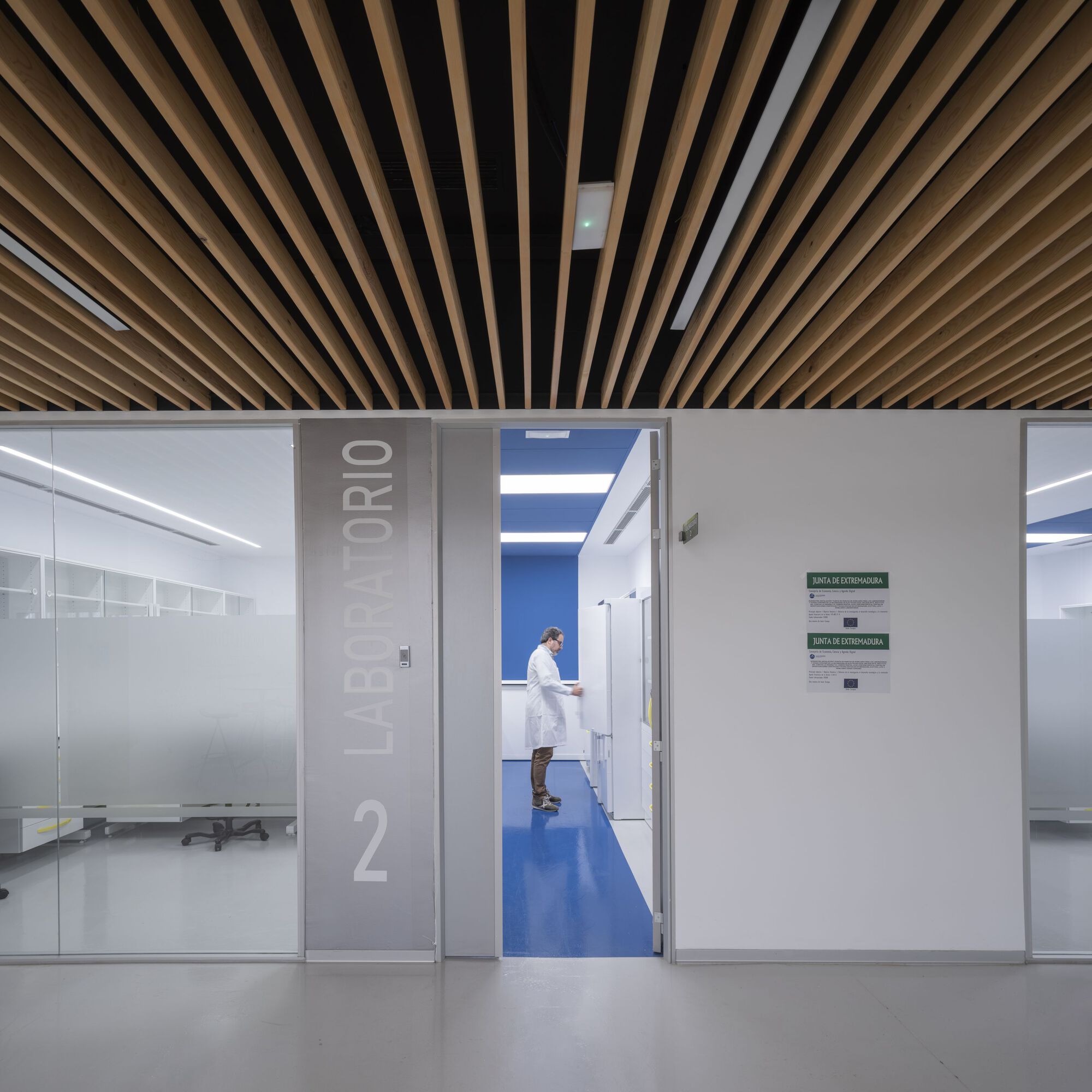 Corridor view through open doorway showing a technician in white coat standing in blue-floored laboratory