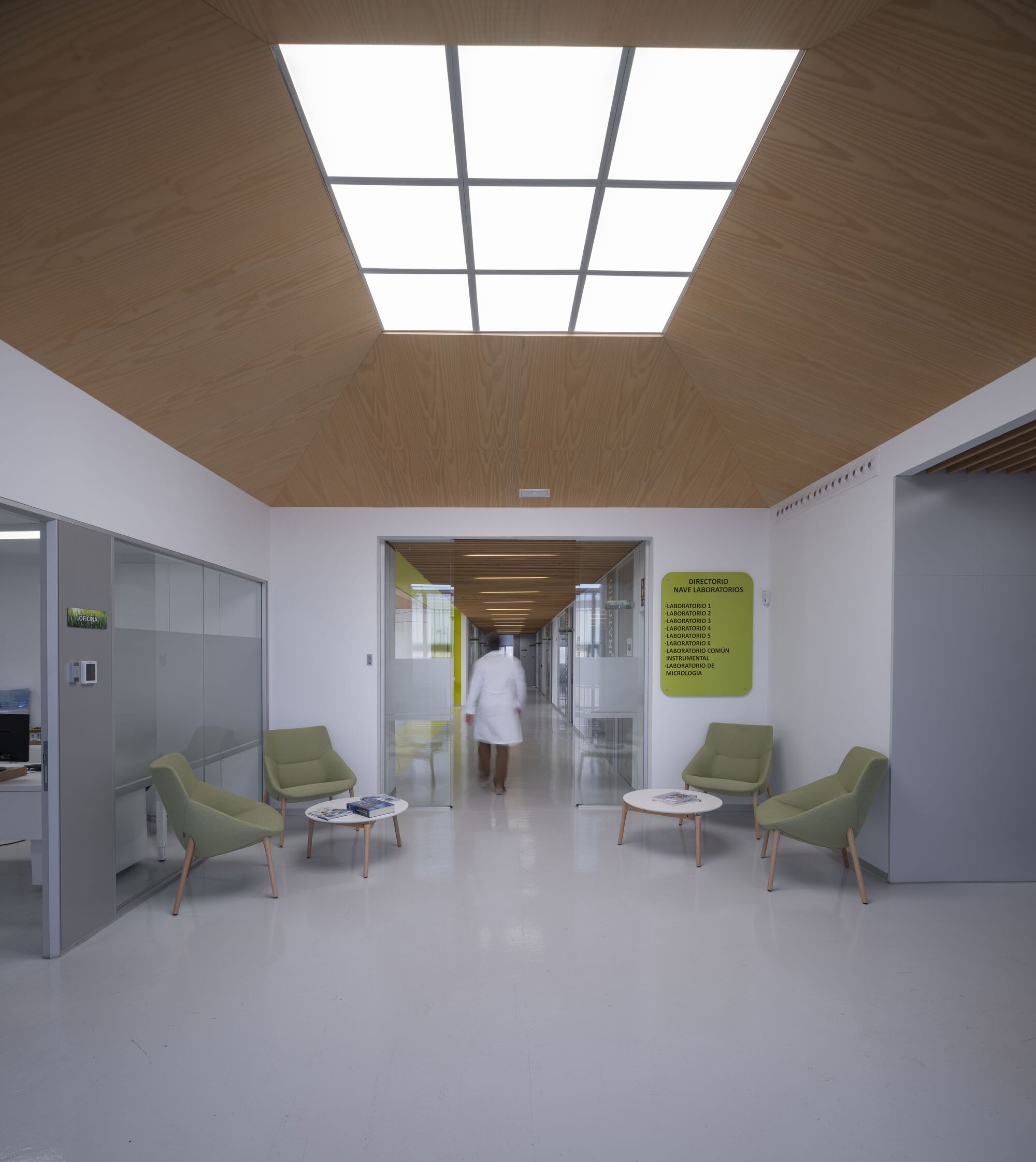 Reception area with timber-clad ceiling skylight, green armchairs and a figure passing through glazed doorway