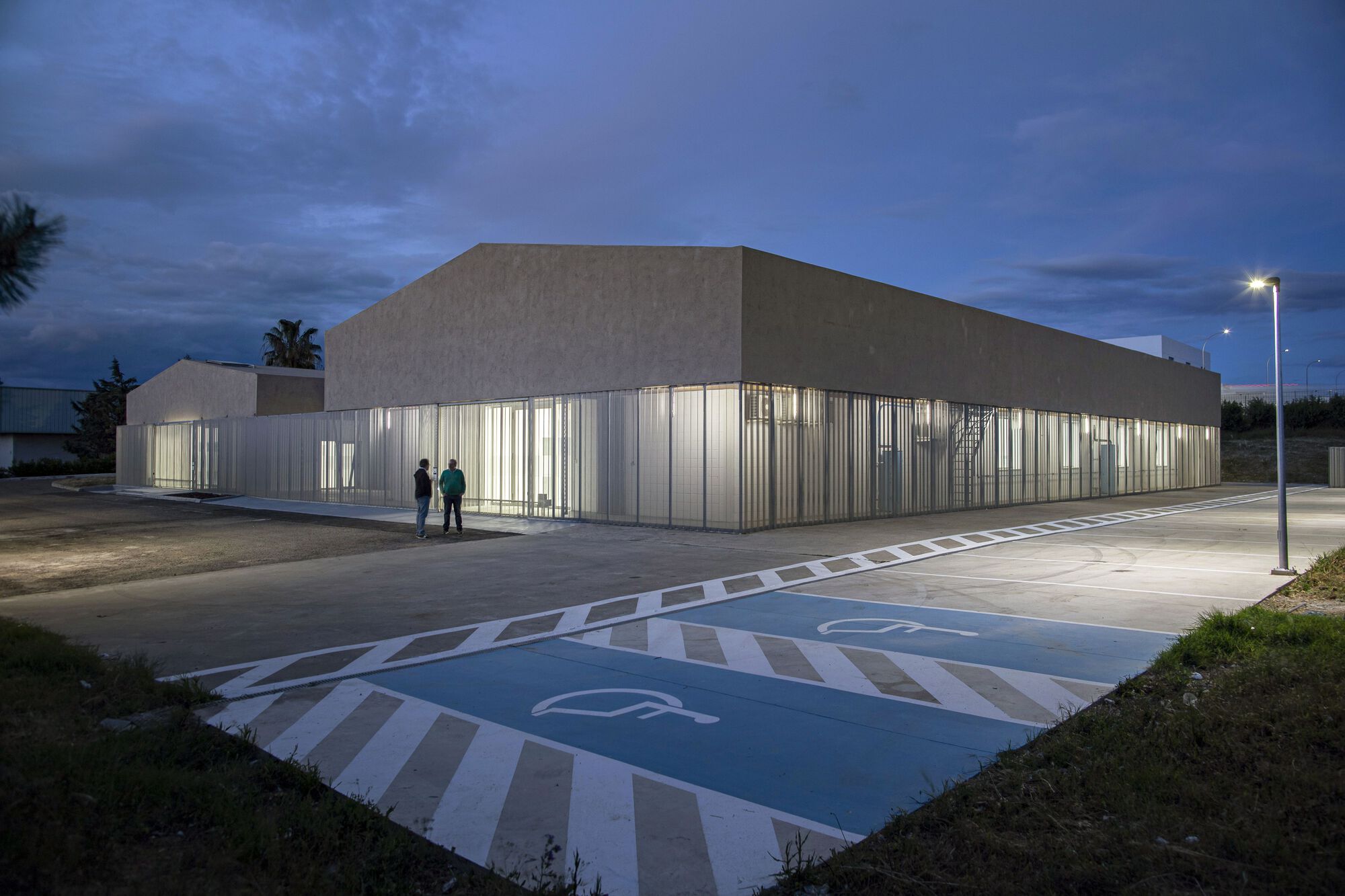 Illuminated translucent corrugated facade wrapping the building corner with two figures standing outside at twilight