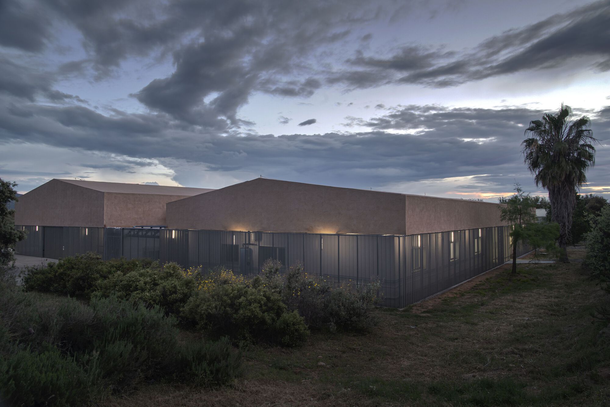 Backlit corrugated metal cladding beneath tan stucco volumes with native planting and stormy evening skies