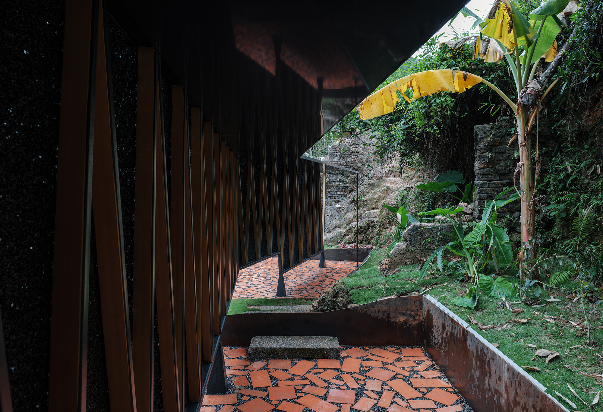 Covered walkway with timber slat screen and broken red terracotta paving leading to a hillside with banana plants