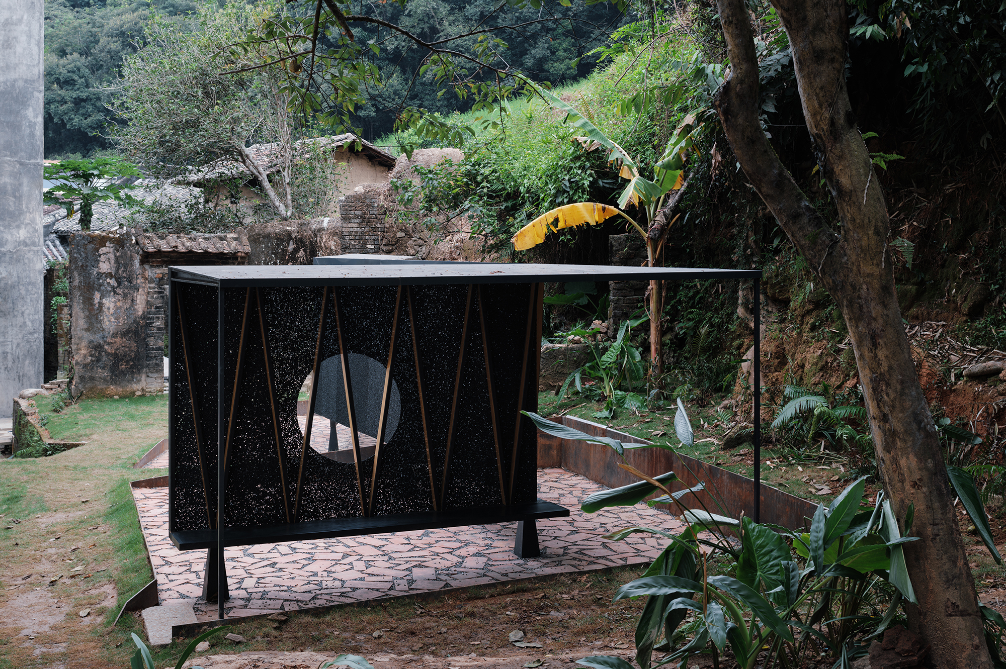 Pavilion with perforated black metal panels and circular opening set among banana trees and stone retaining walls