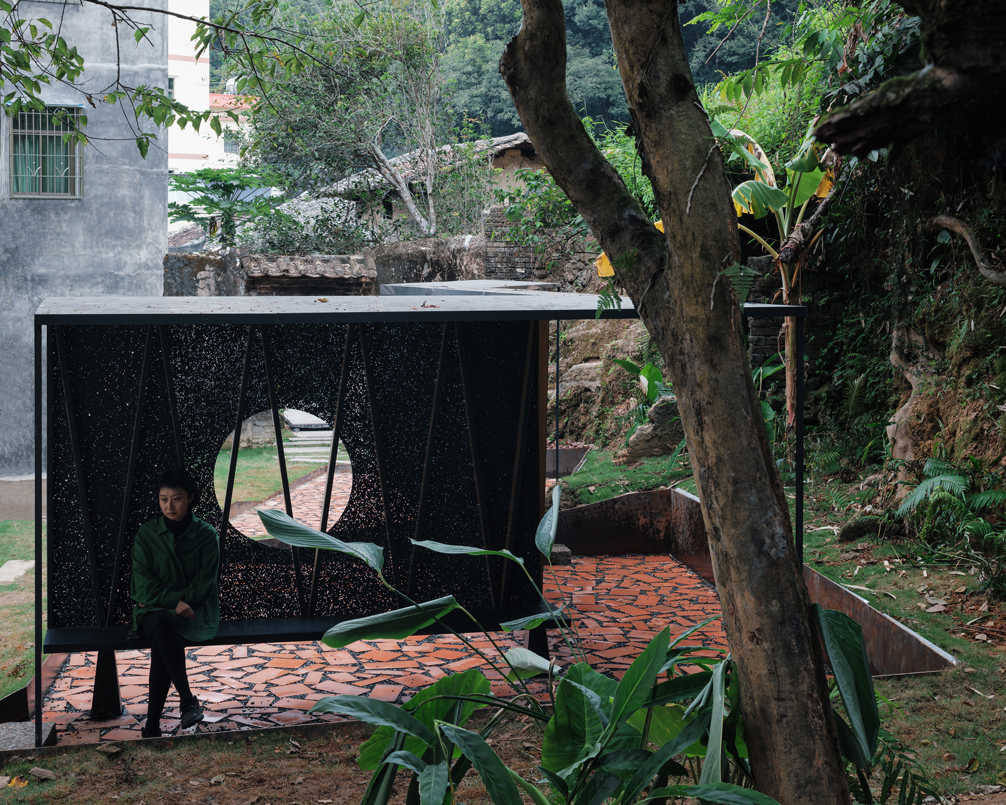 Perforated metal pavilion with circular portal and person seated inside beneath overhanging tree branches