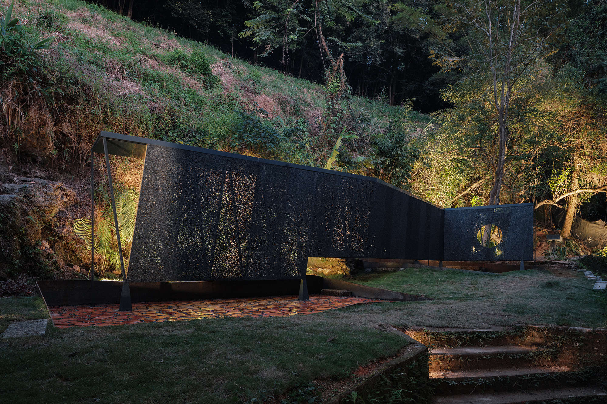 Perforated black pavilion at dusk with interior lighting glowing against forested hillside