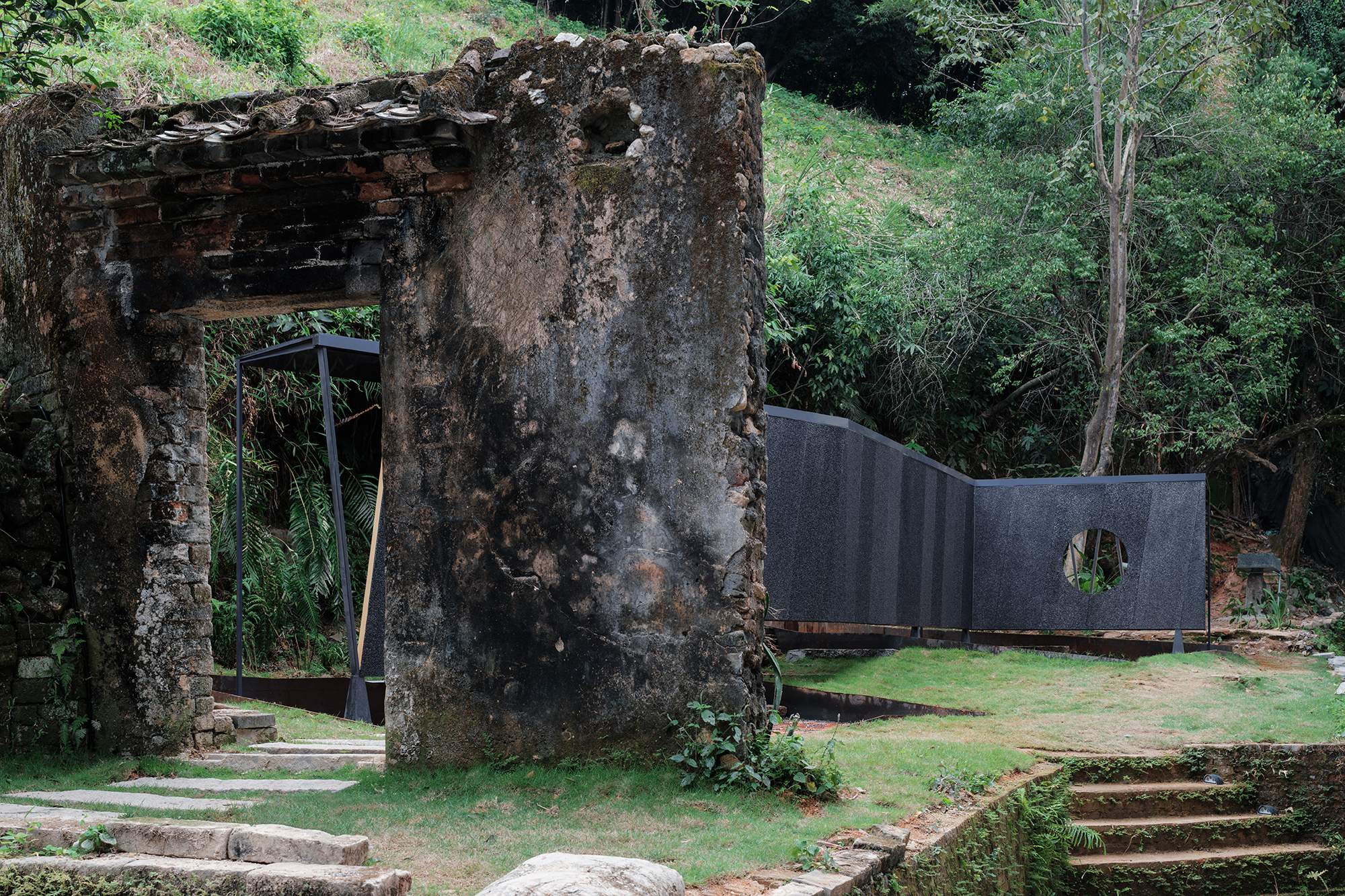 Weathered concrete ruins adjacent to dark curved wall with circular aperture in grass clearing