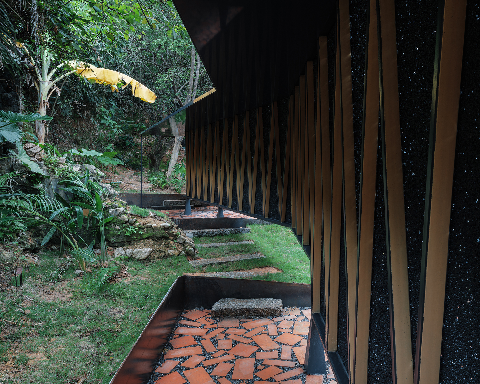 Curved terrace with red brick paving and vertical timber screen beside stone steps and banana trees