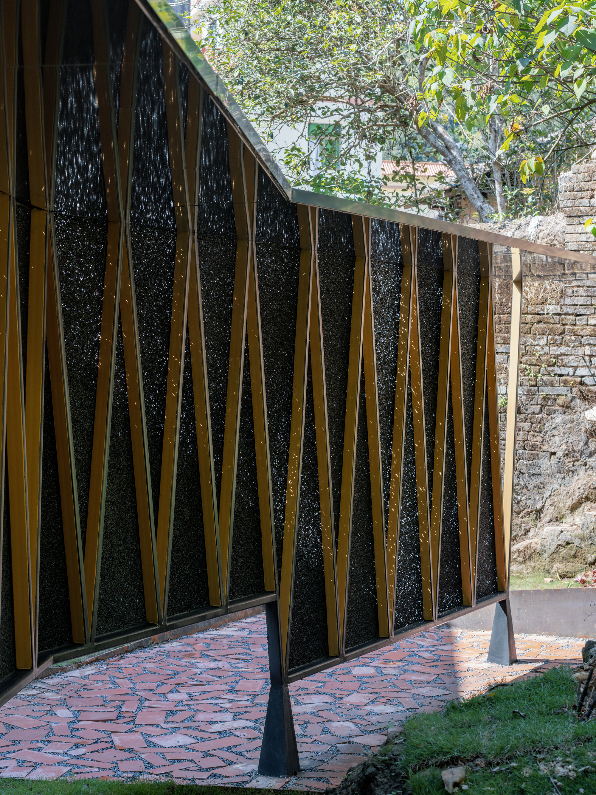 Perforated metal screen wall with diagonal bronze framing above a terracotta paver terrace beneath overhanging trees