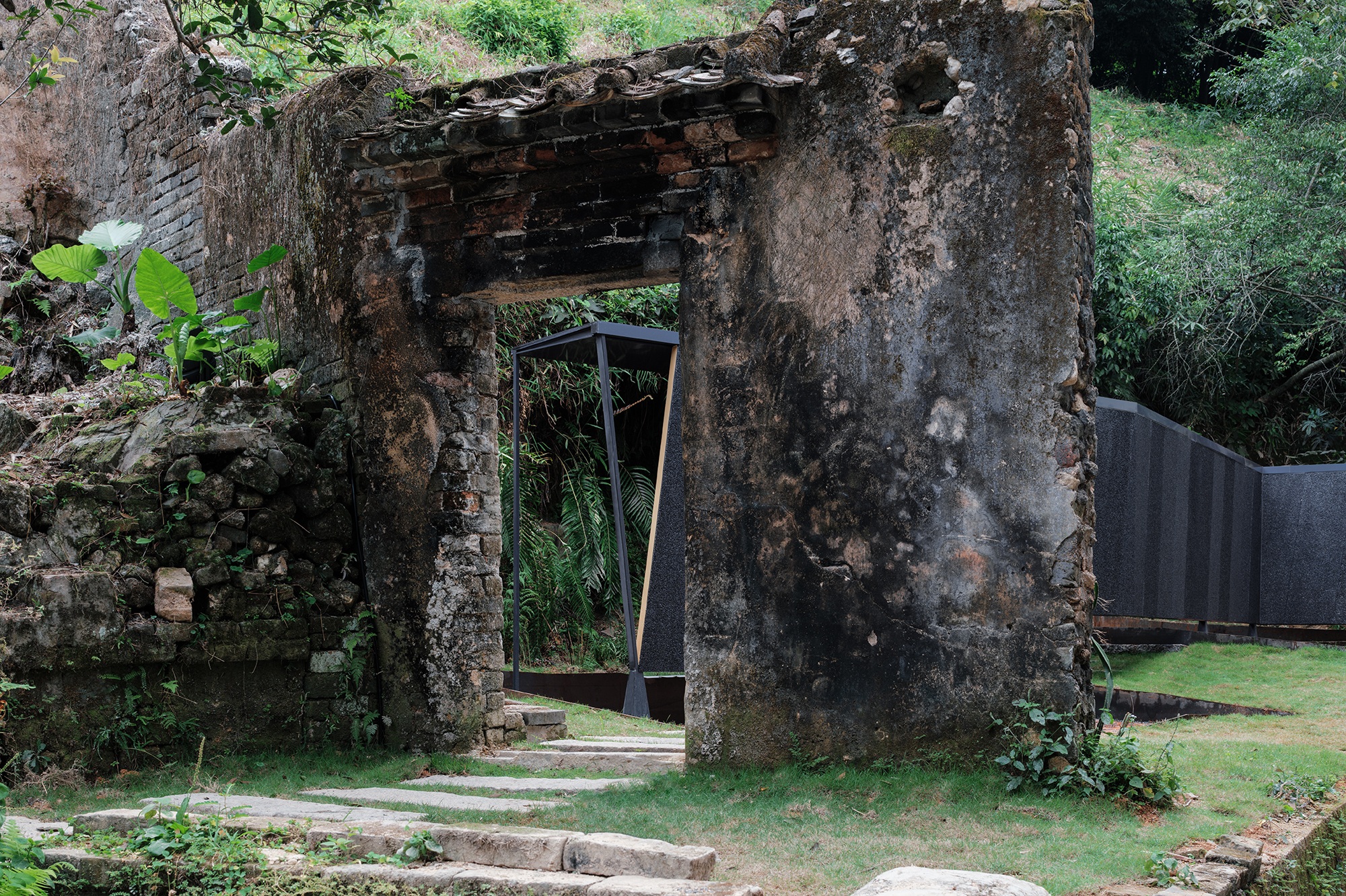 Crumbling stone ruin with a metal-framed door opening looking onto planted lawn and tropical growth