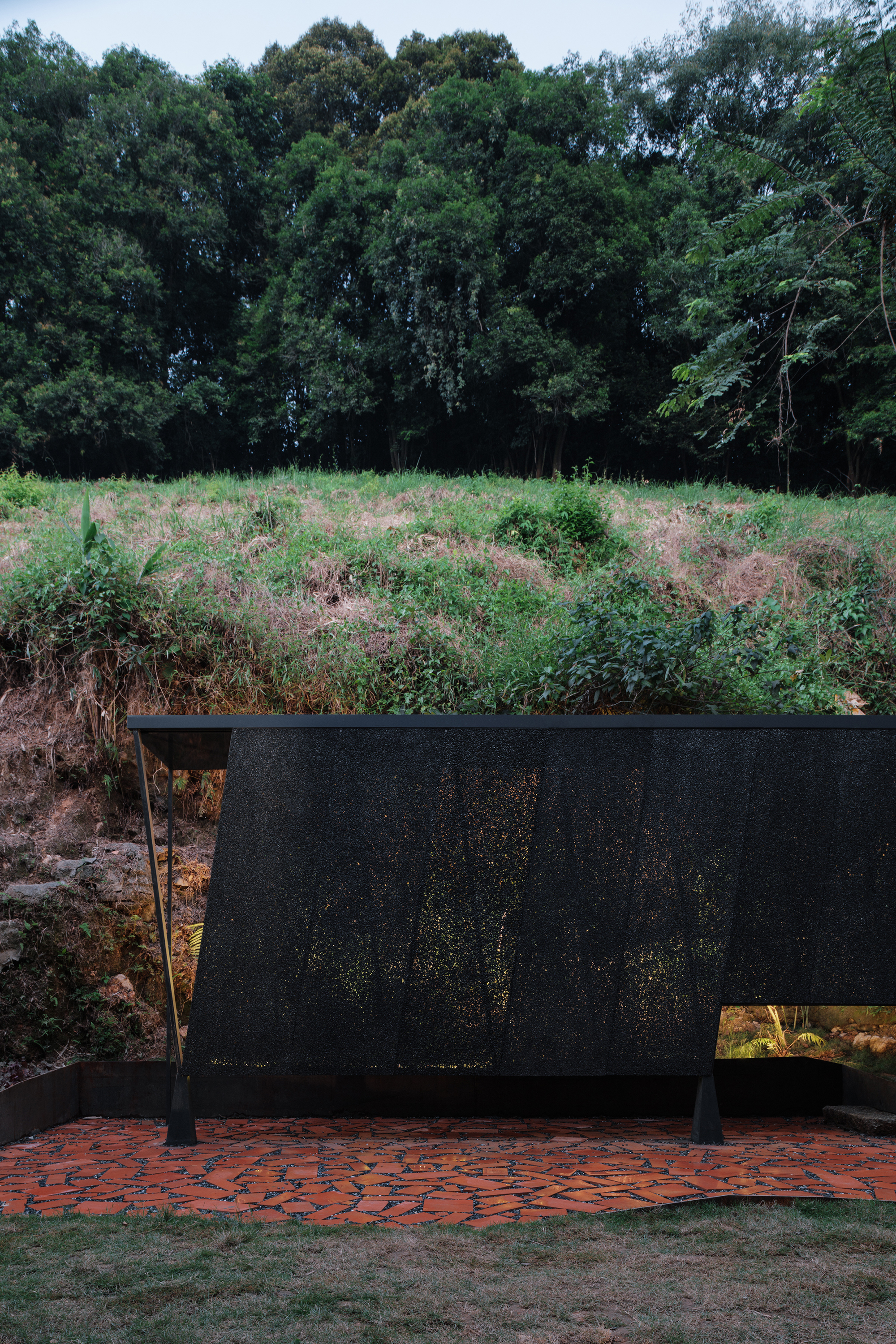 Freestanding perforated black screen wall on a herringbone brick terrace facing an overgrown hillside