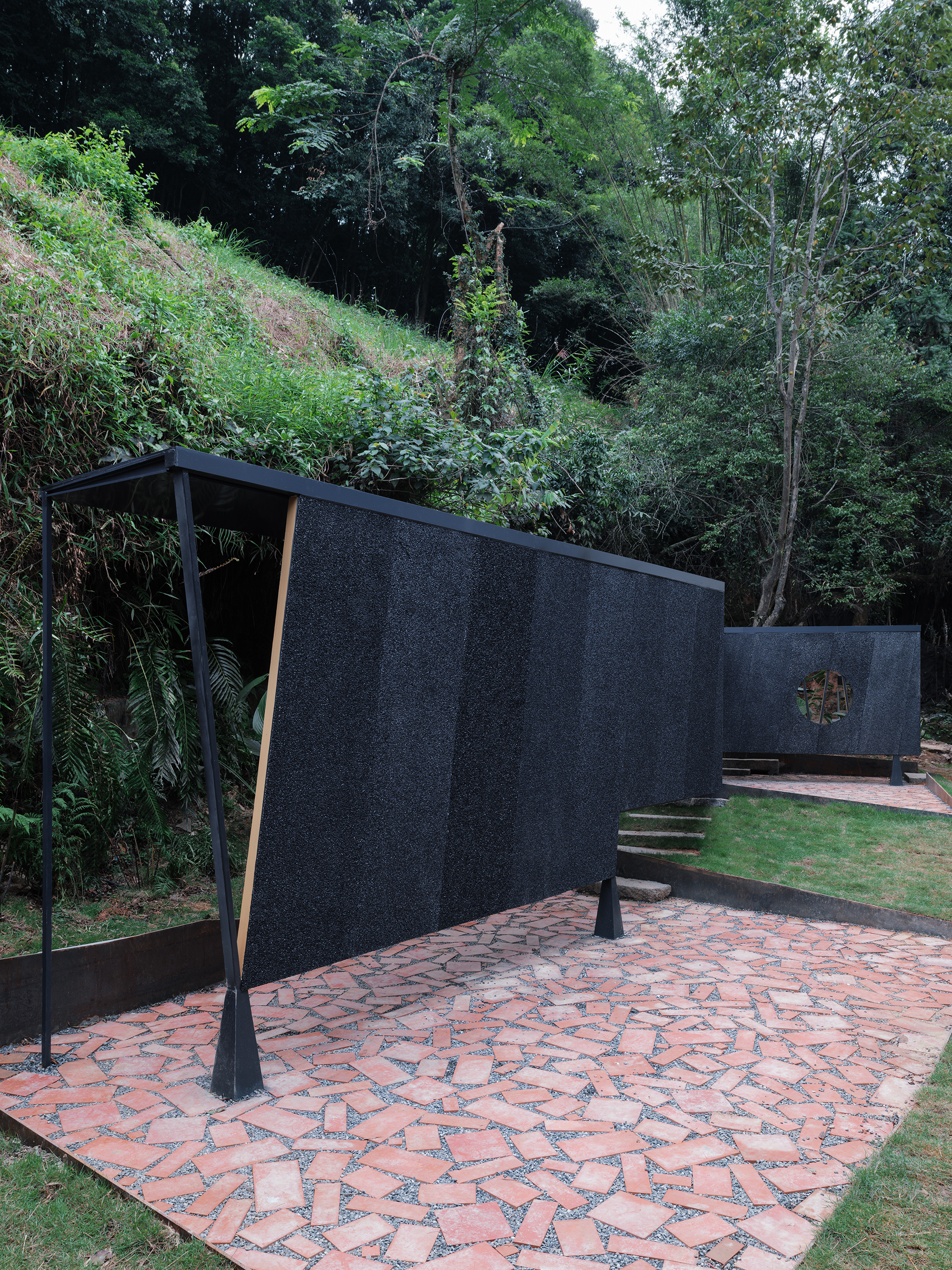 Angular screen wall with angled support struts on a patterned brick terrace backed by forested slope