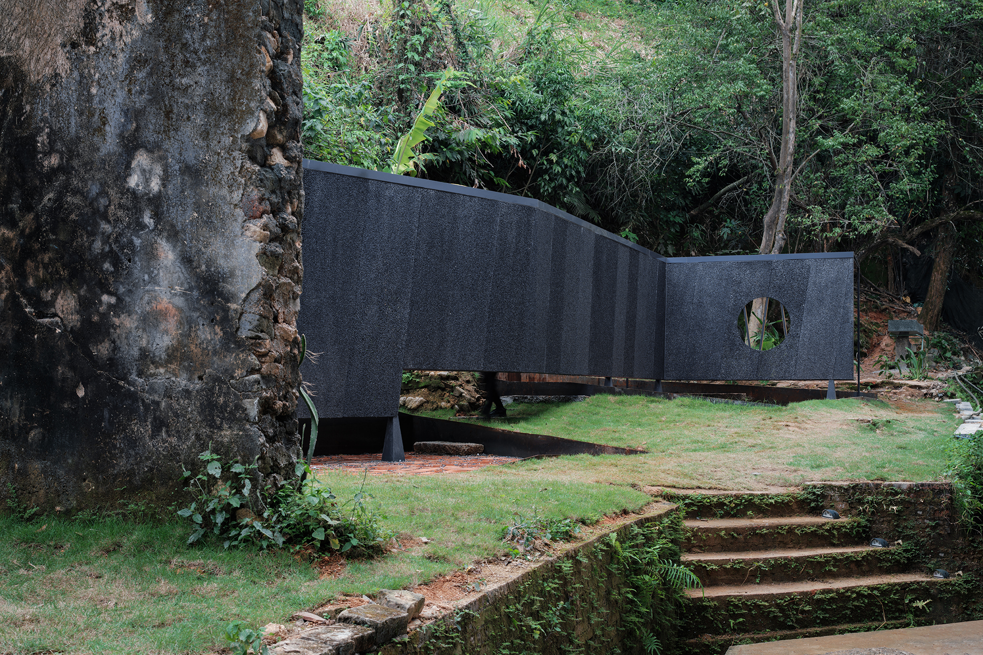 Curving black textured wall with circular opening set against terraced lawn and stone steps beneath dense foliage