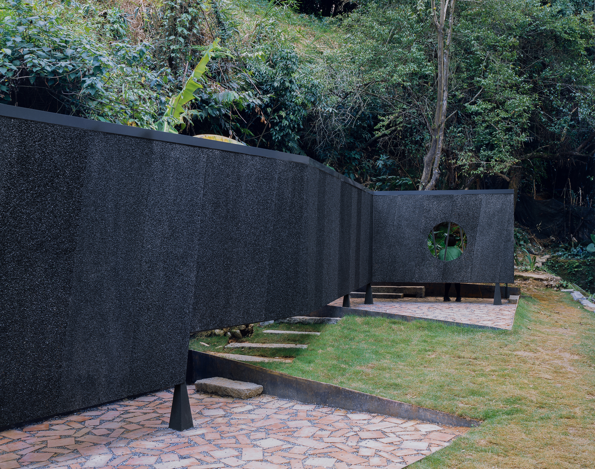 Paired curving black walls with circular aperture creating courtyard spaces connected by patterned stone paving