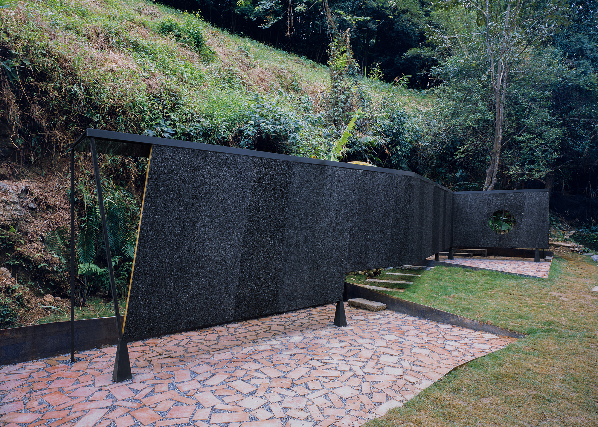 Black textured wall curving through the site with patterned paving terraces and sloped planted banks beyond