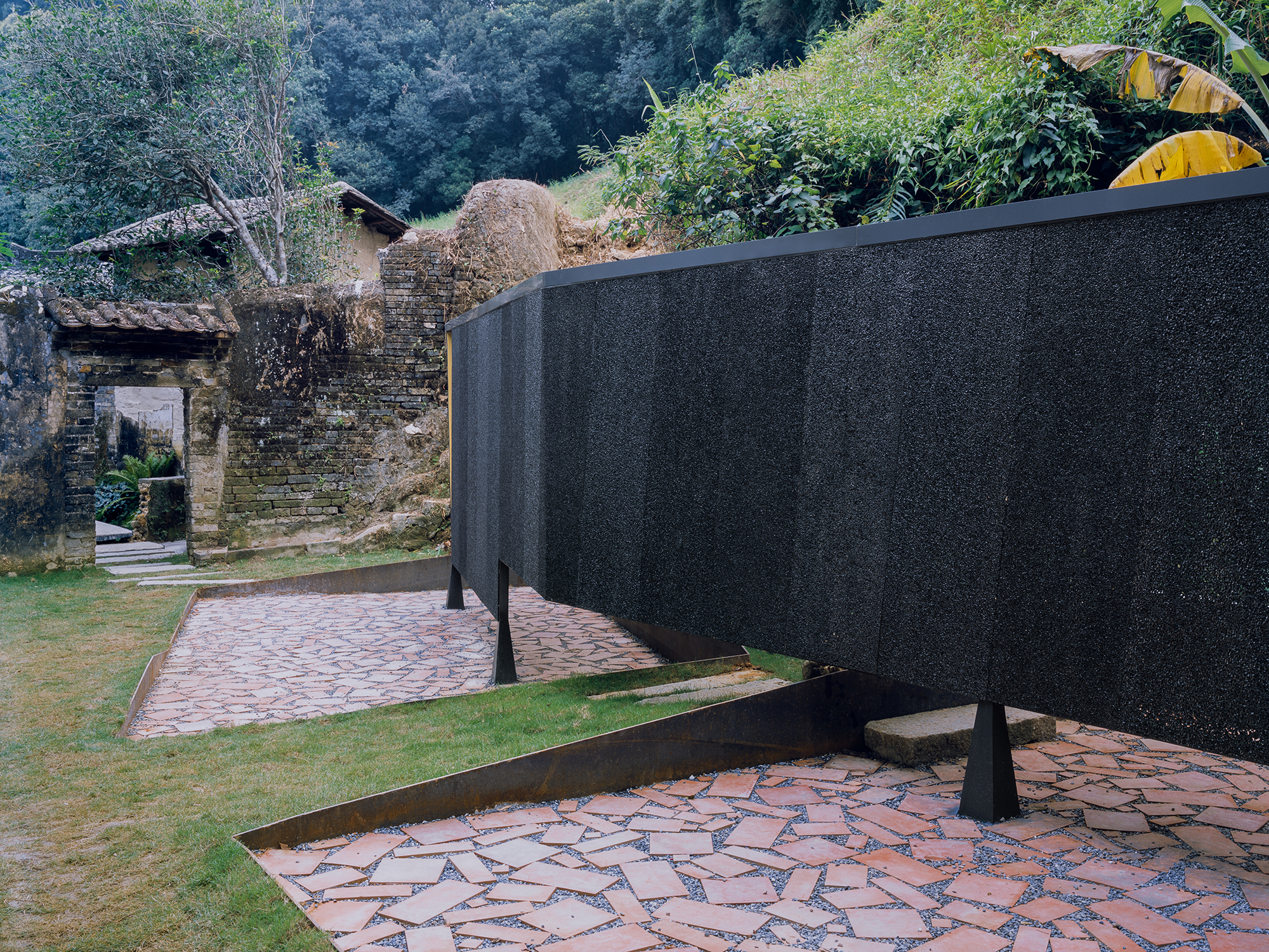 Curving black wall bisecting paved terraces with weathered stone ruins visible on the hillside behind