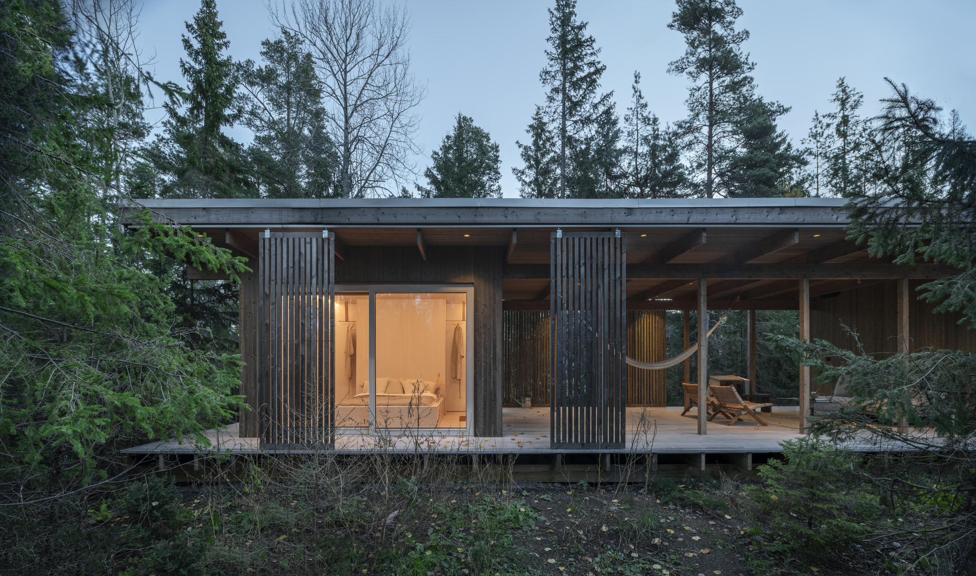 Flat-roofed timber cabin with vertical slatted screens and glazed openings nestled among evergreen trees at dusk