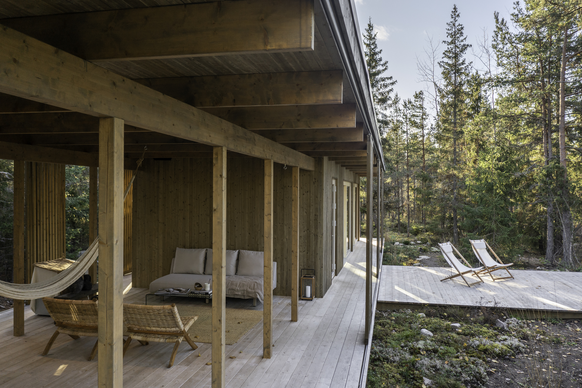 Covered timber porch with exposed columns and beams opening to a forest deck with lounge chairs