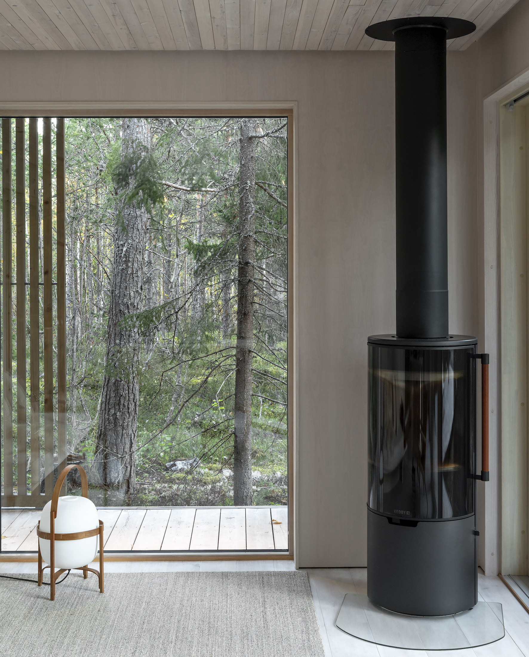 Freestanding black cylindrical wood stove beside a full-height window overlooking the forest