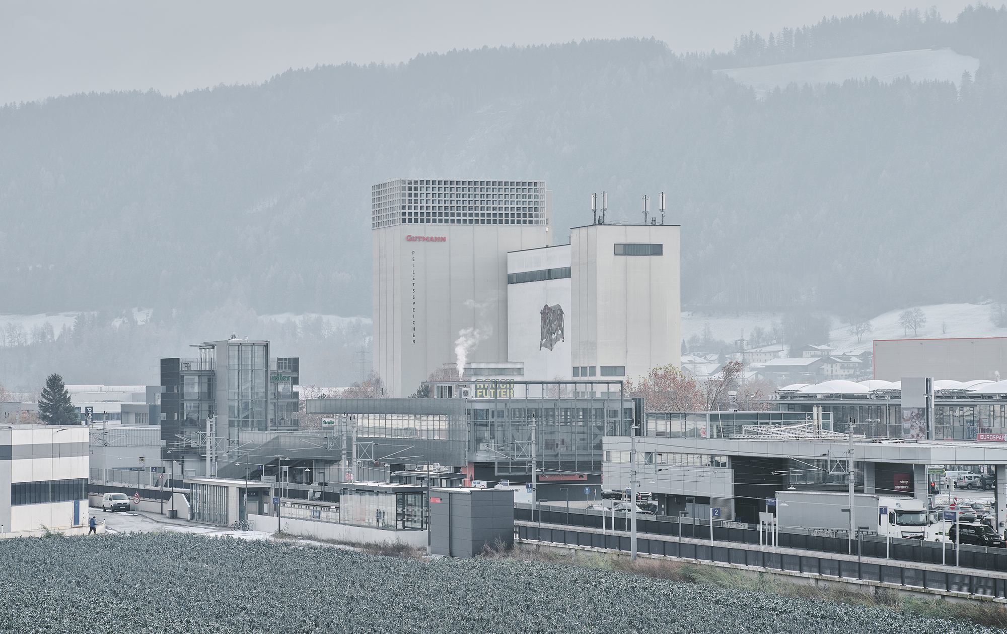 Industrial complex with concrete tower and perforated parapet set against misty forested hills in winter
