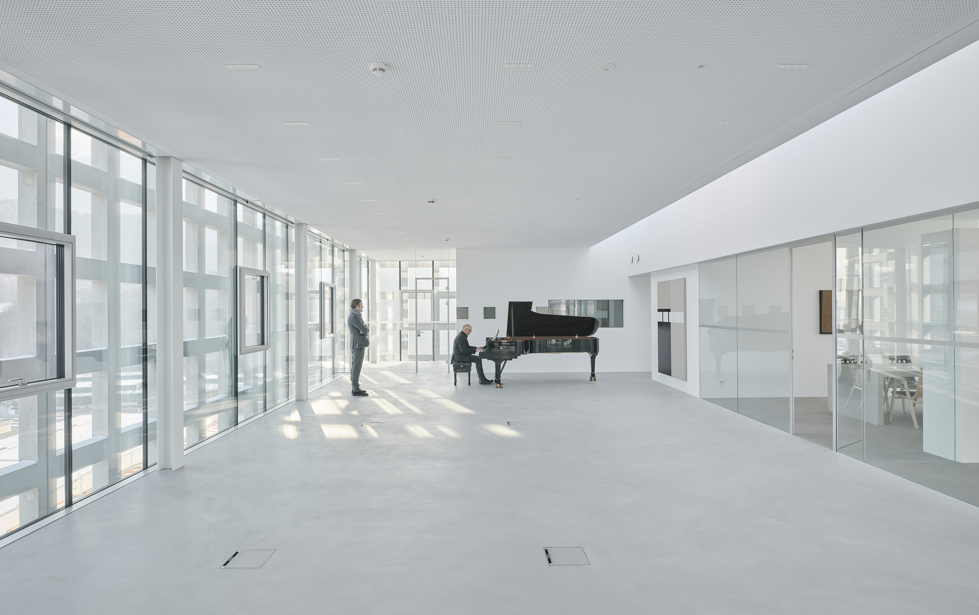 Open white interior with floor-to-ceiling glazing, a grand piano and two people in soft daylight
