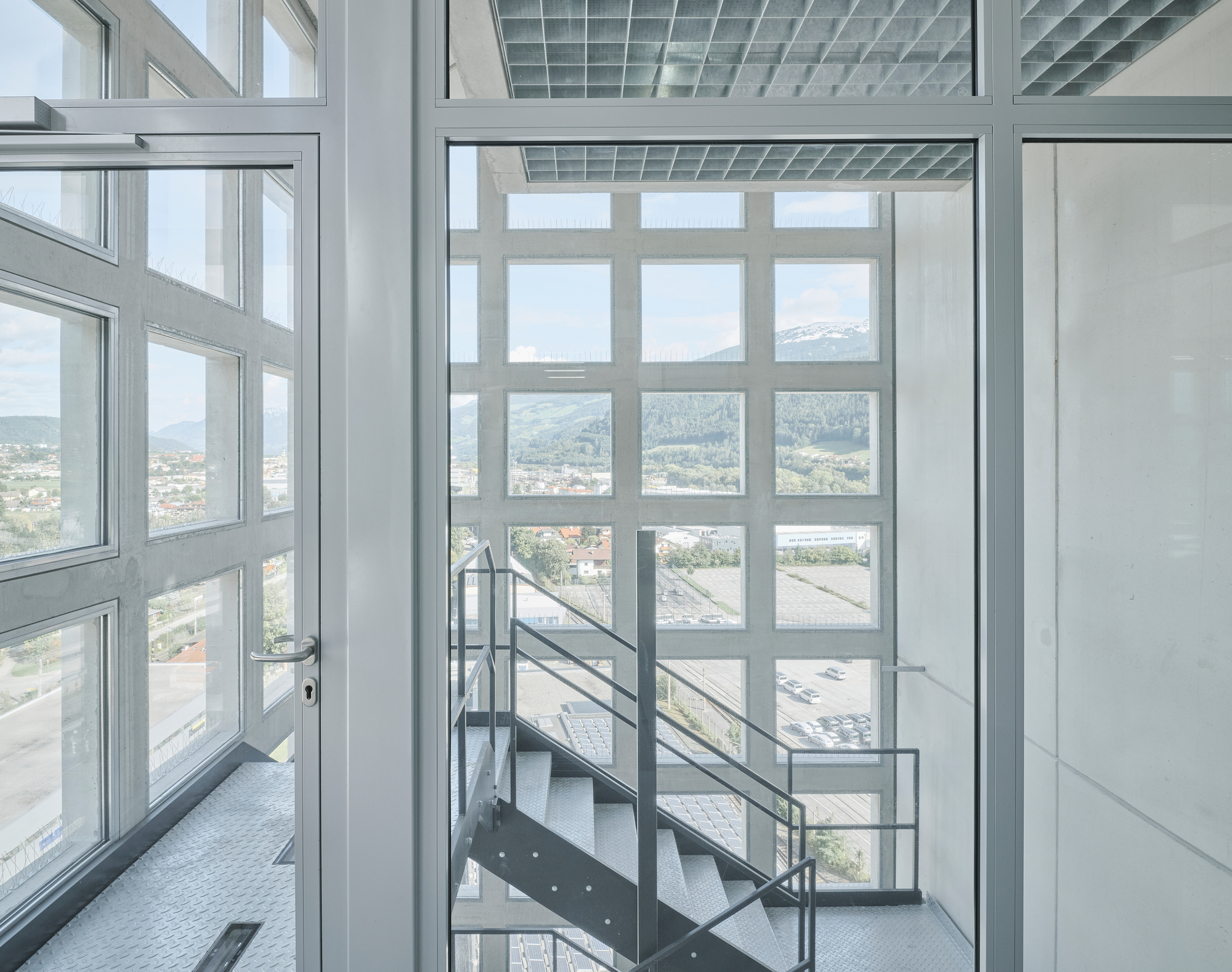 Steel staircase with wire mesh railing rising through a white-framed glass curtain wall overlooking distant mountains