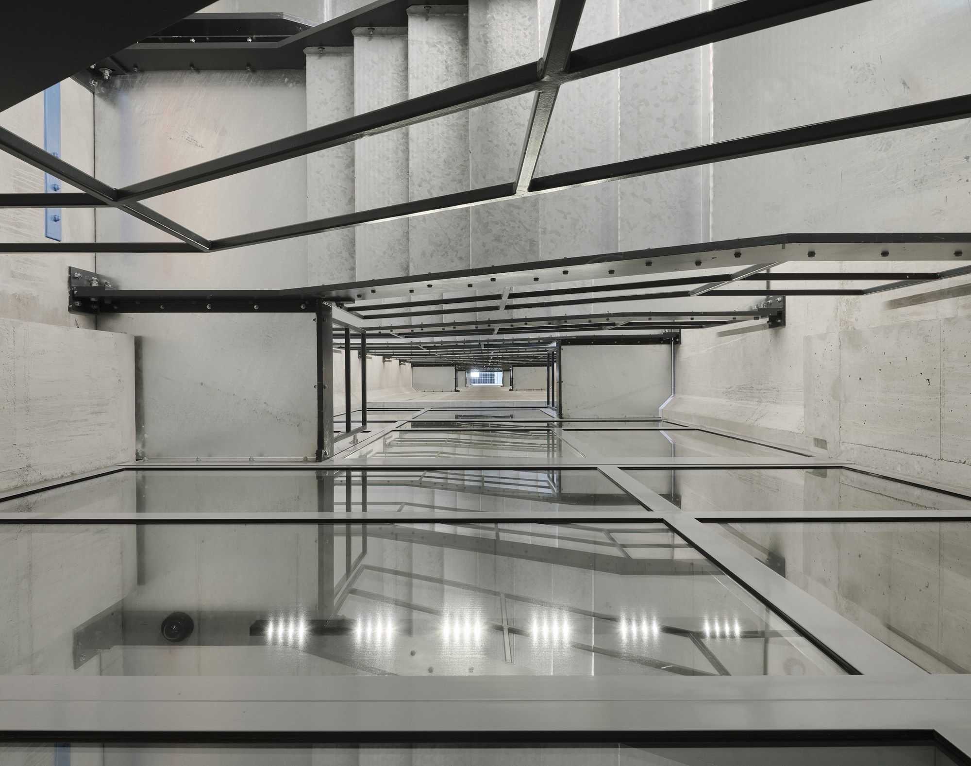 Looking down through a concrete elevator shaft with steel beams and reflective glass floor panels