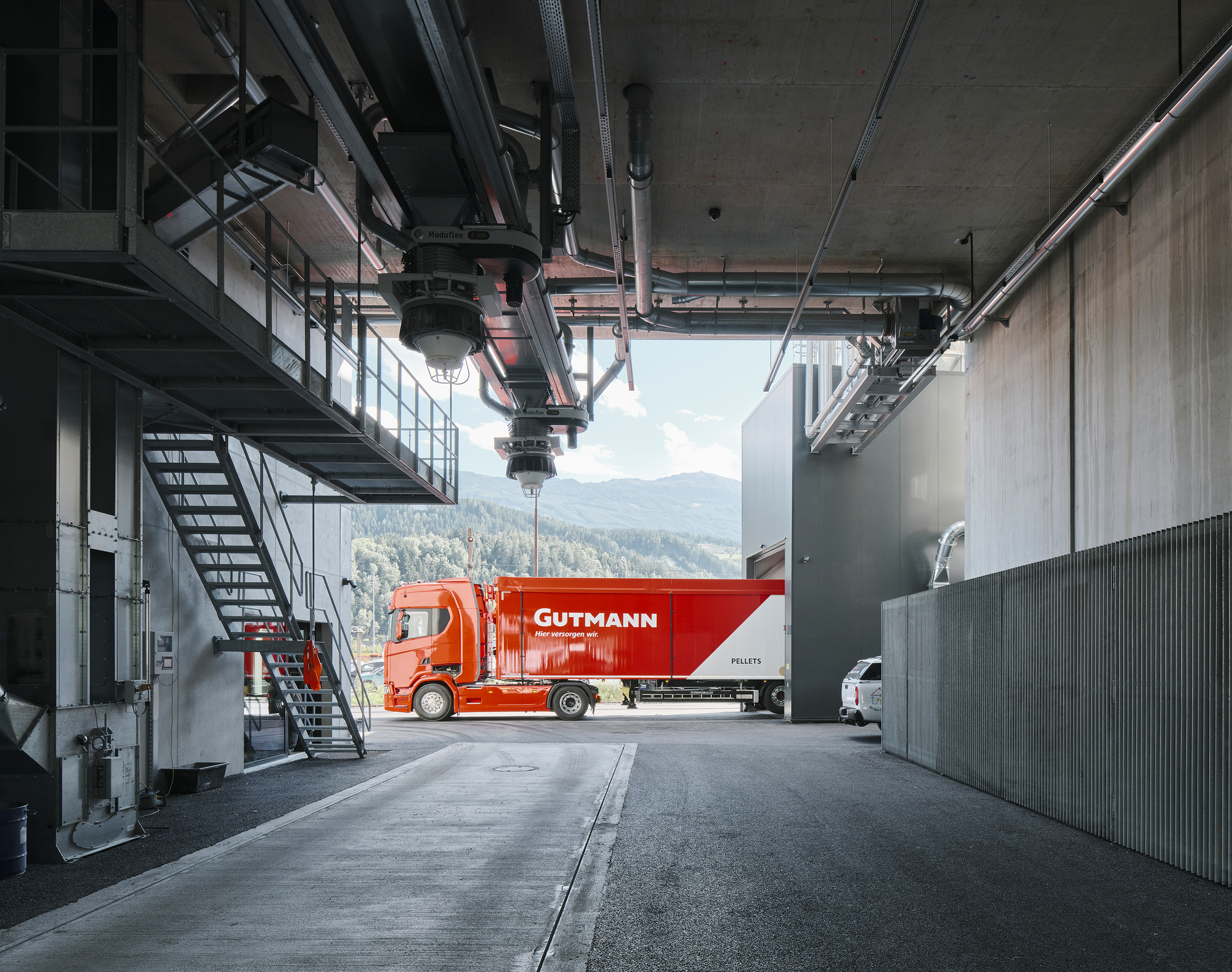 Covered loading bay with exposed concrete ceiling, steel walkway, and red delivery truck framed by mountain view