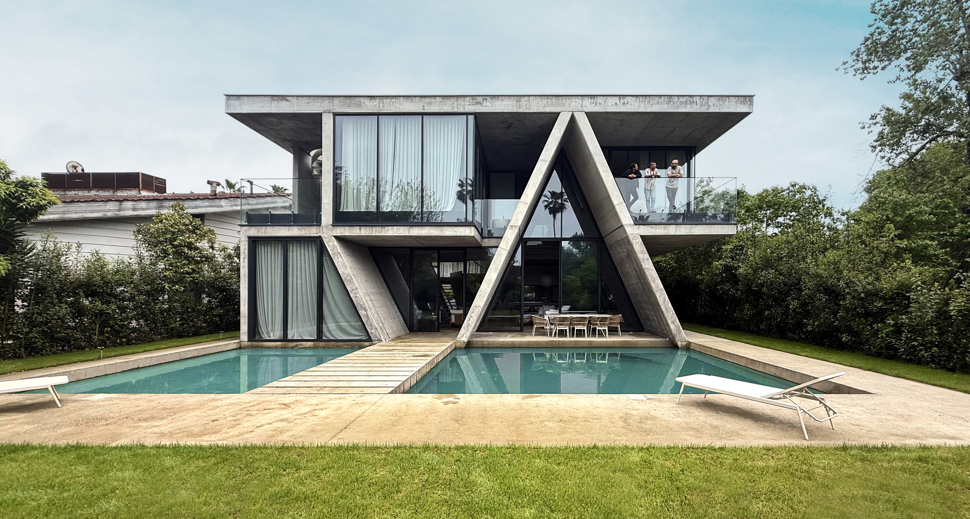 Garden facade with angled concrete supports framing a swimming pool and timber deck under overcast sky
