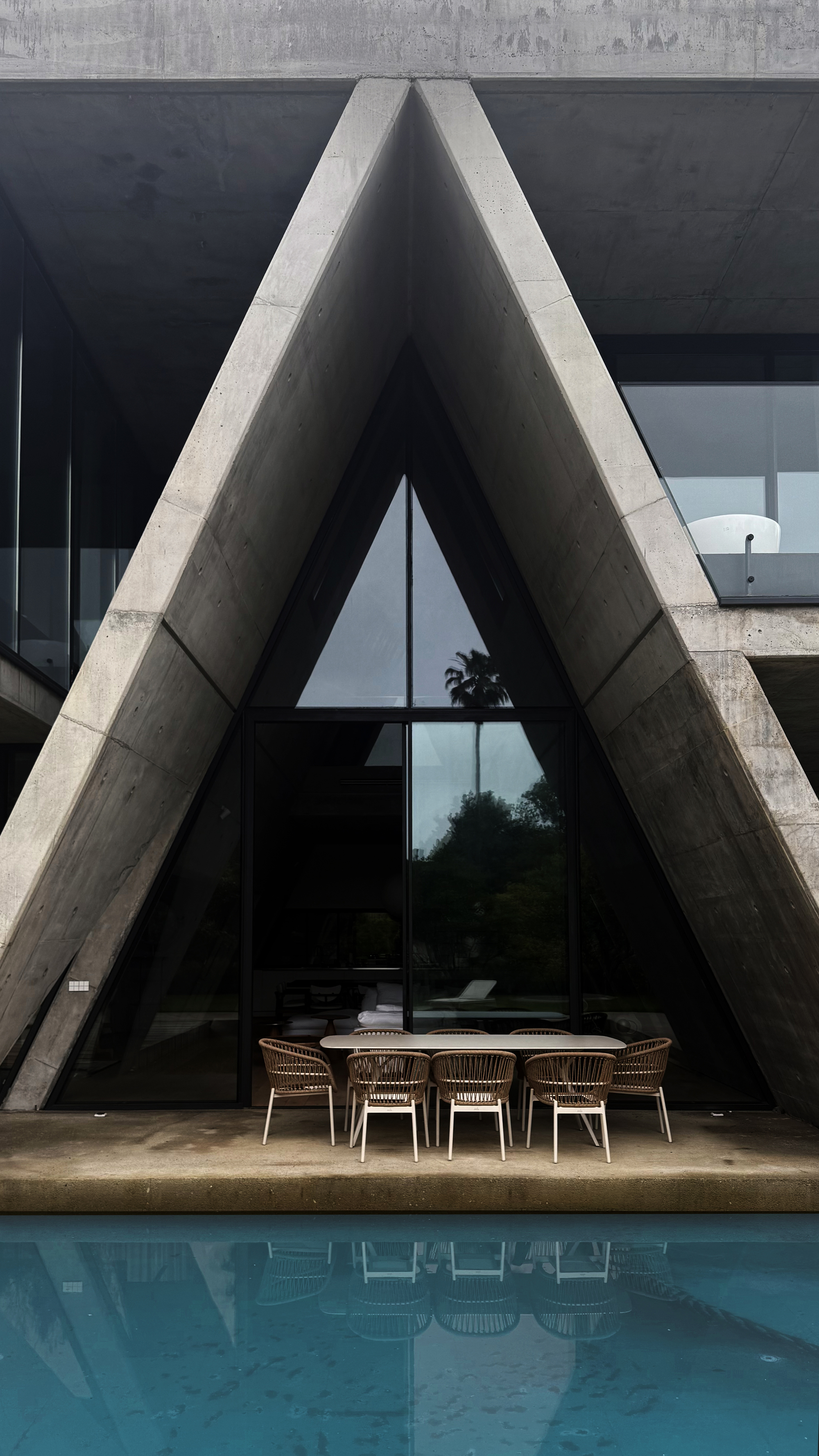 View of the triangular concrete portal framing the glazed entrance with a dining table beside the pool