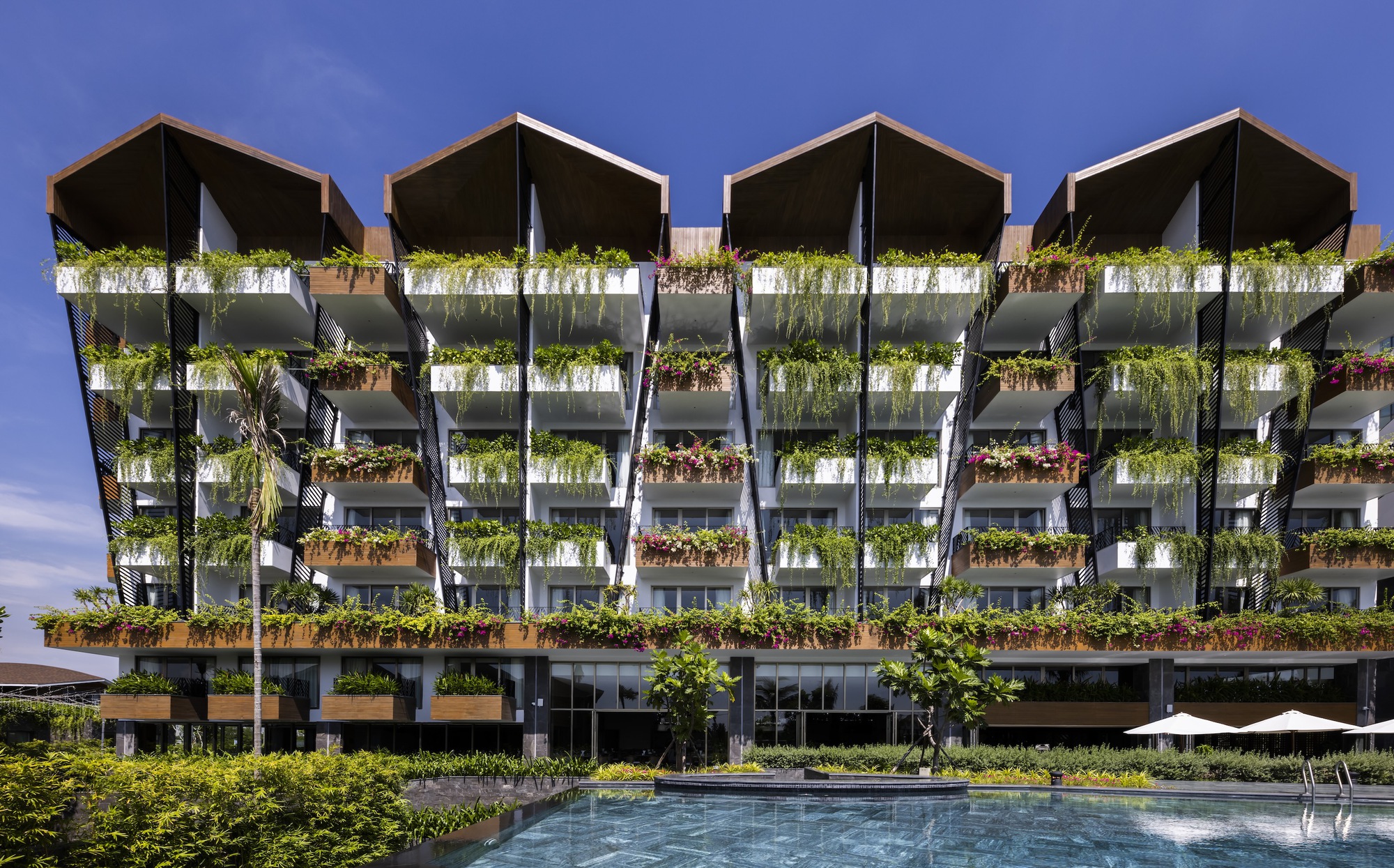 Facade with planted balconies and dark canopy roofs viewed across a swimming pool under blue sky