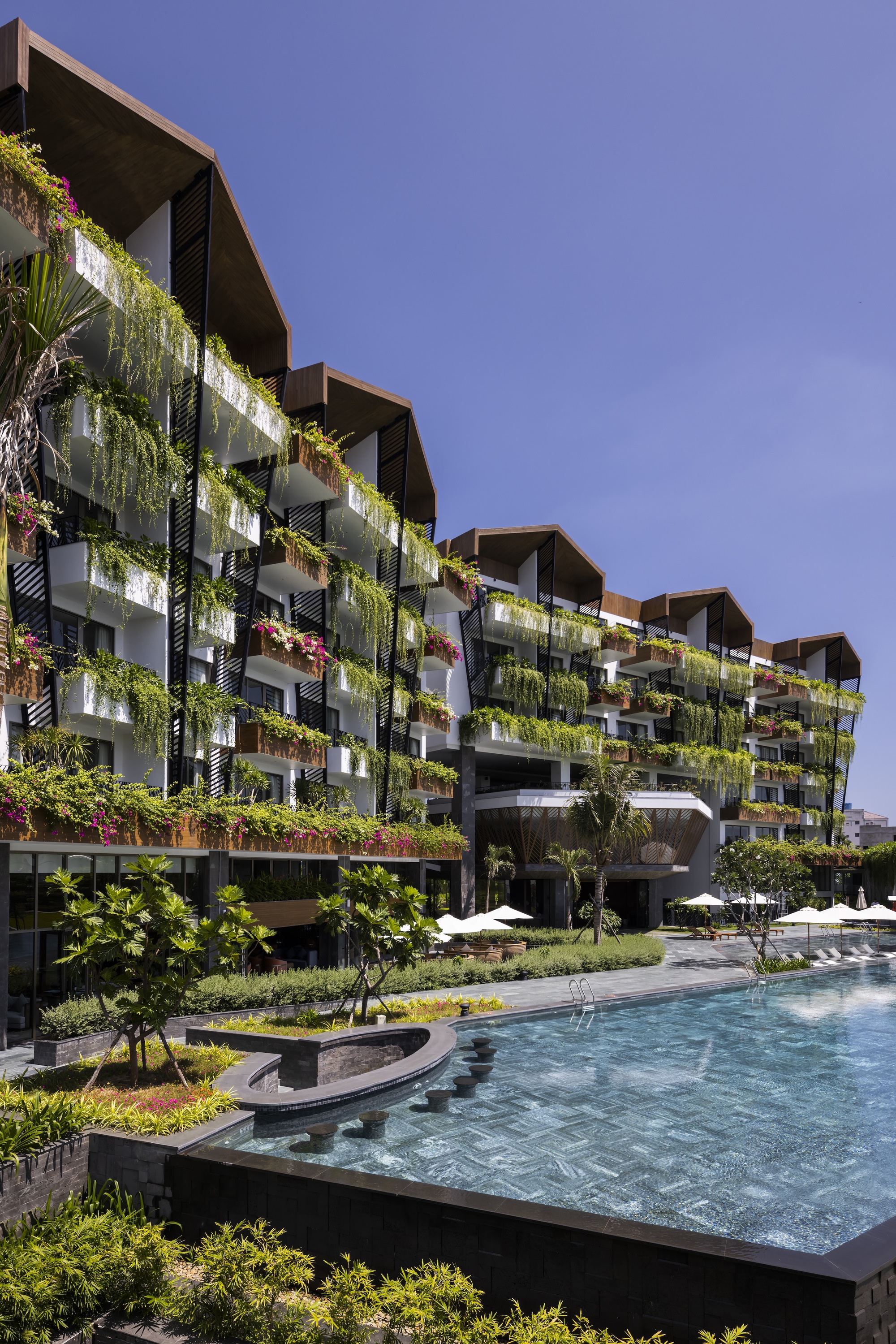 Planted facade rising above curved pool edge and walkway with young trees in afternoon light